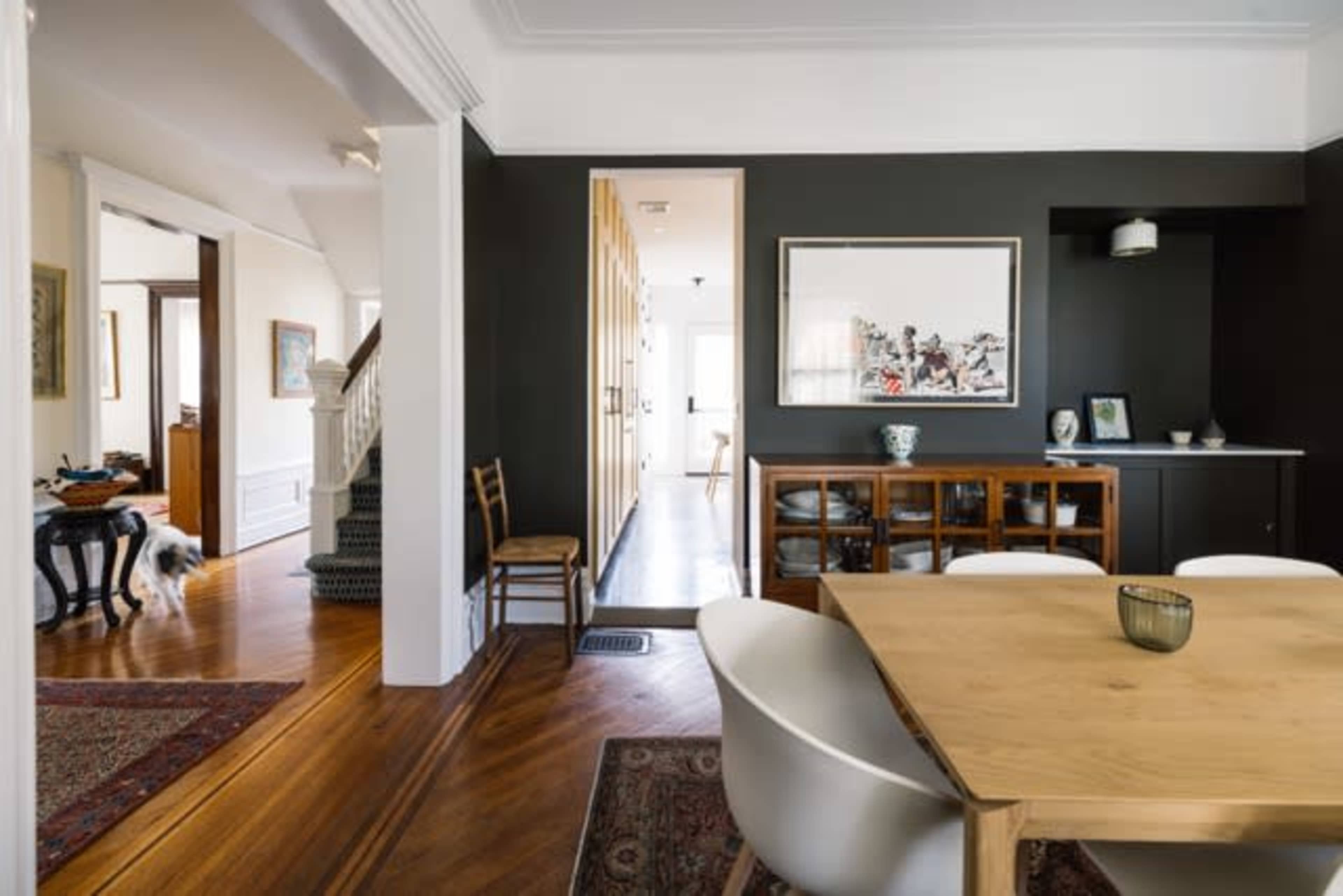 The image shows a modern dining room with a wooden table and white chairs, leading to a hallway that connects to other parts of a house with dark walls and wooden accents.