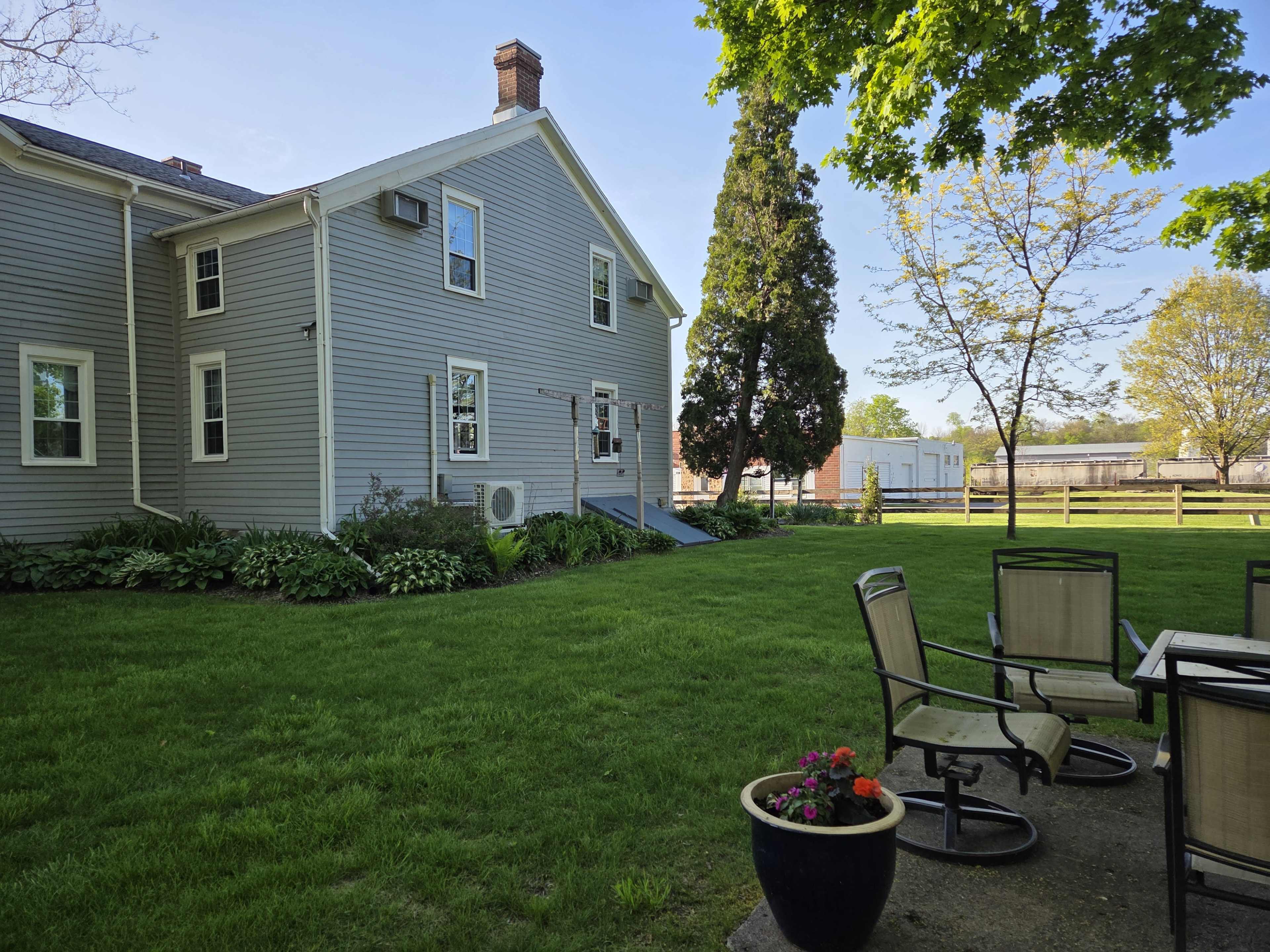 The image shows a grassy backyard with a house on one side, a patio set with chairs, and a pathway leading to a visible trailer in the background.