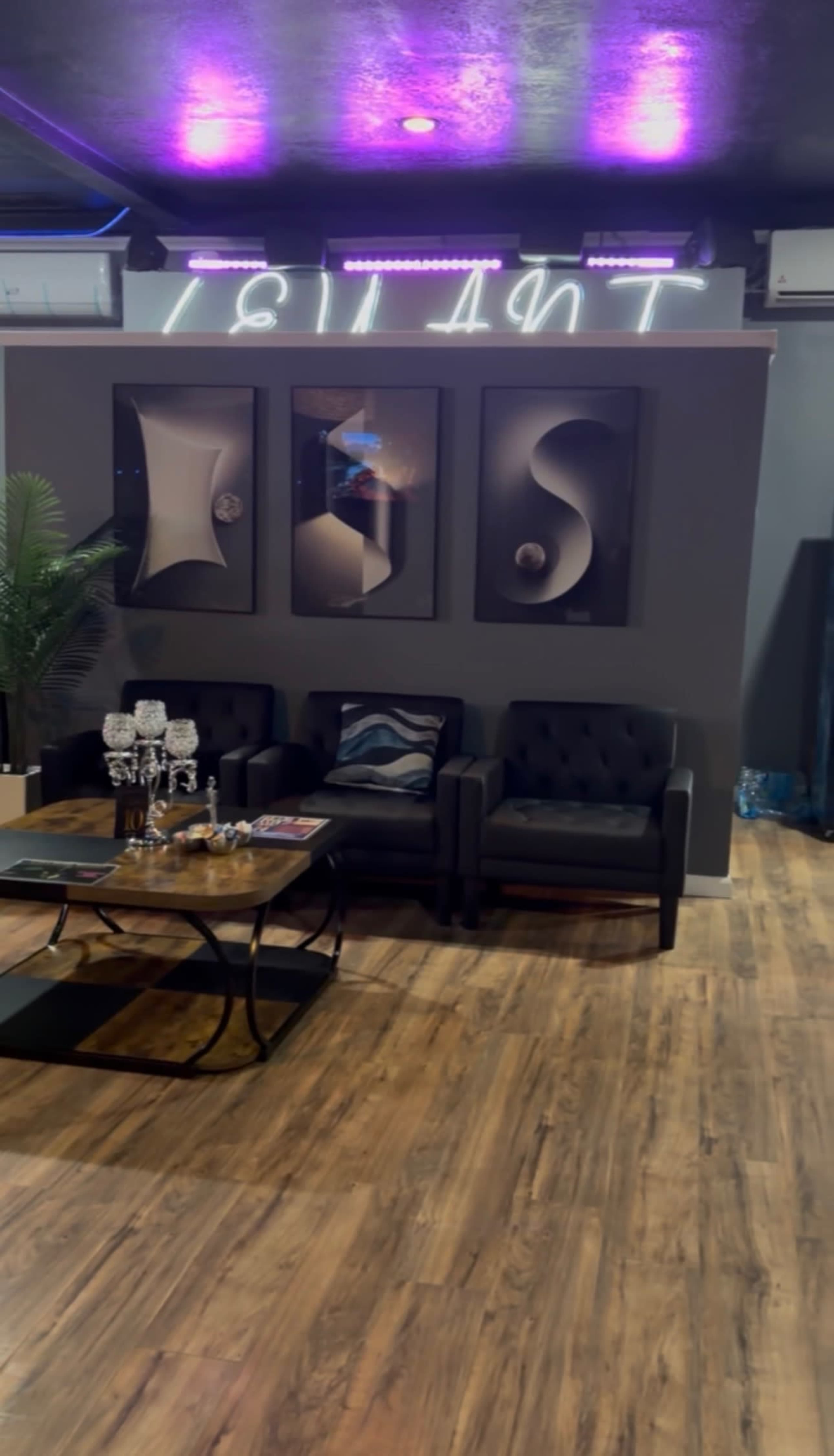 The image shows a modern seating area with two black upholstered chairs and a glass coffee table, set against a gray wall adorned with three abstract framed artworks.