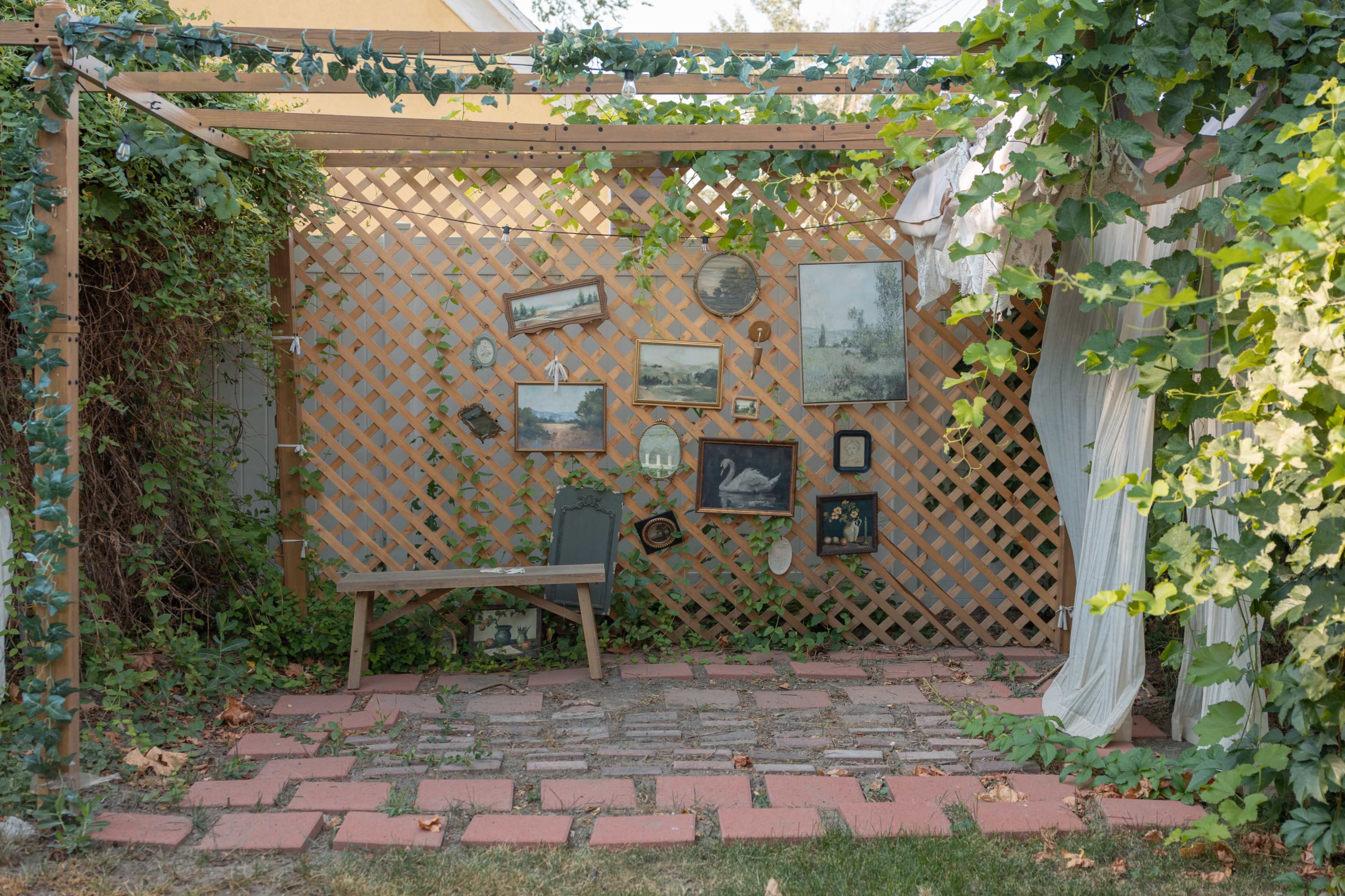 A small outdoor space features a wooden lattice structure adorned with framed pictures and surrounded by greenery and brick paving.