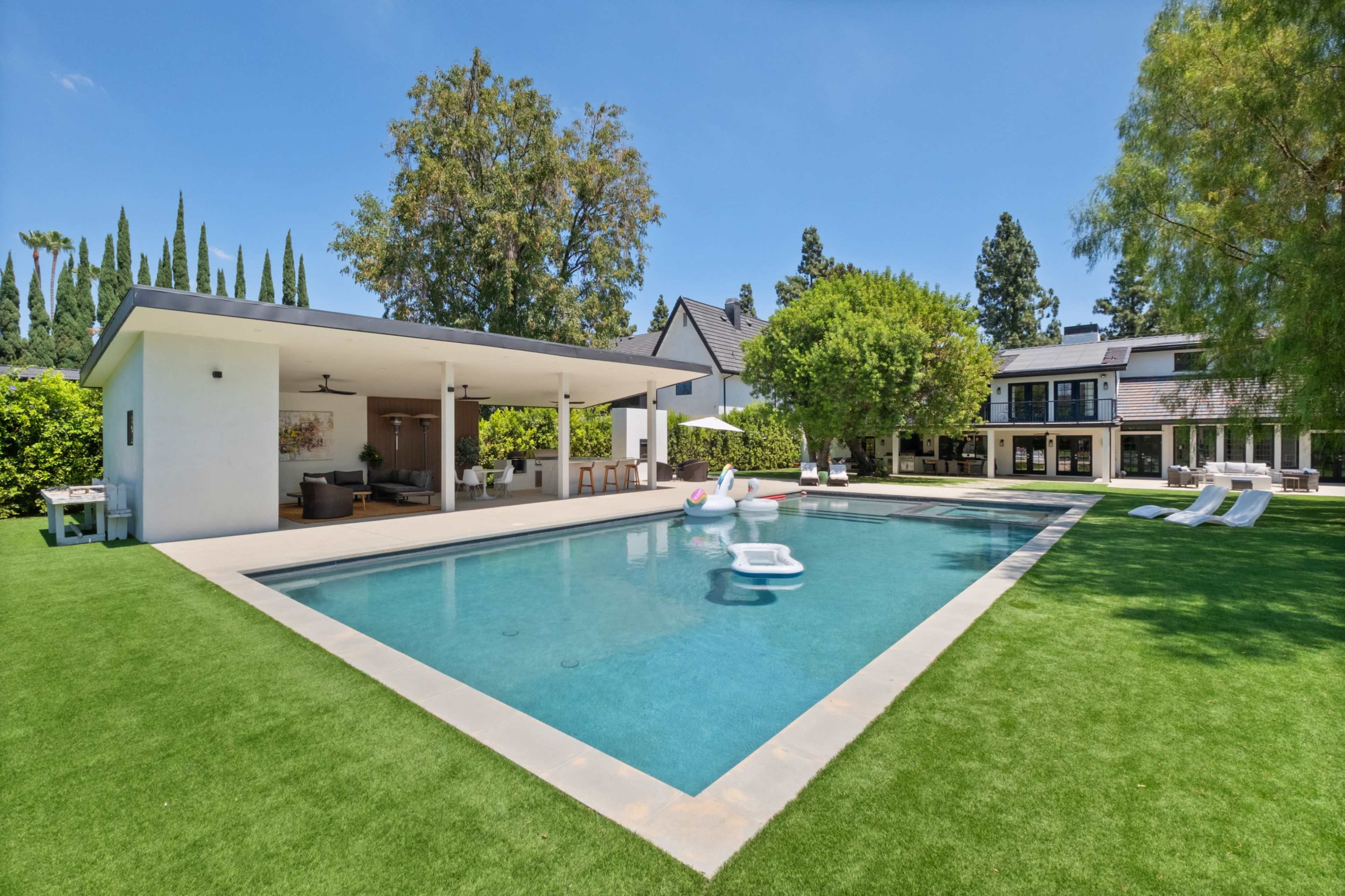 The image features a modern backyard with a rectangular swimming pool surrounded by manicured lawns, lounge chairs, and a covered patio area.
