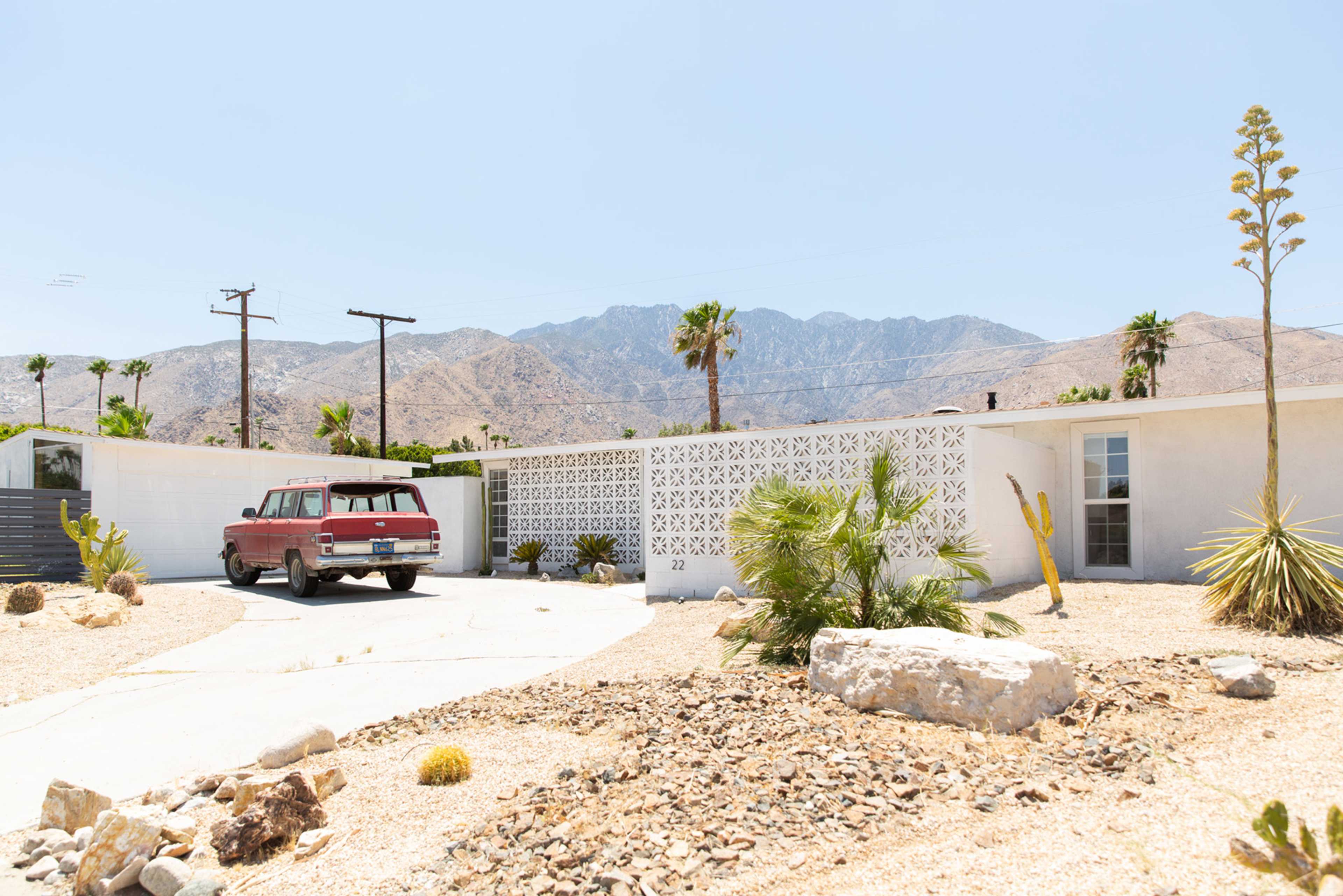 A red pickup truck is parked in the driveway of a mid-century styled house in a desert landscape with mountains in the background.