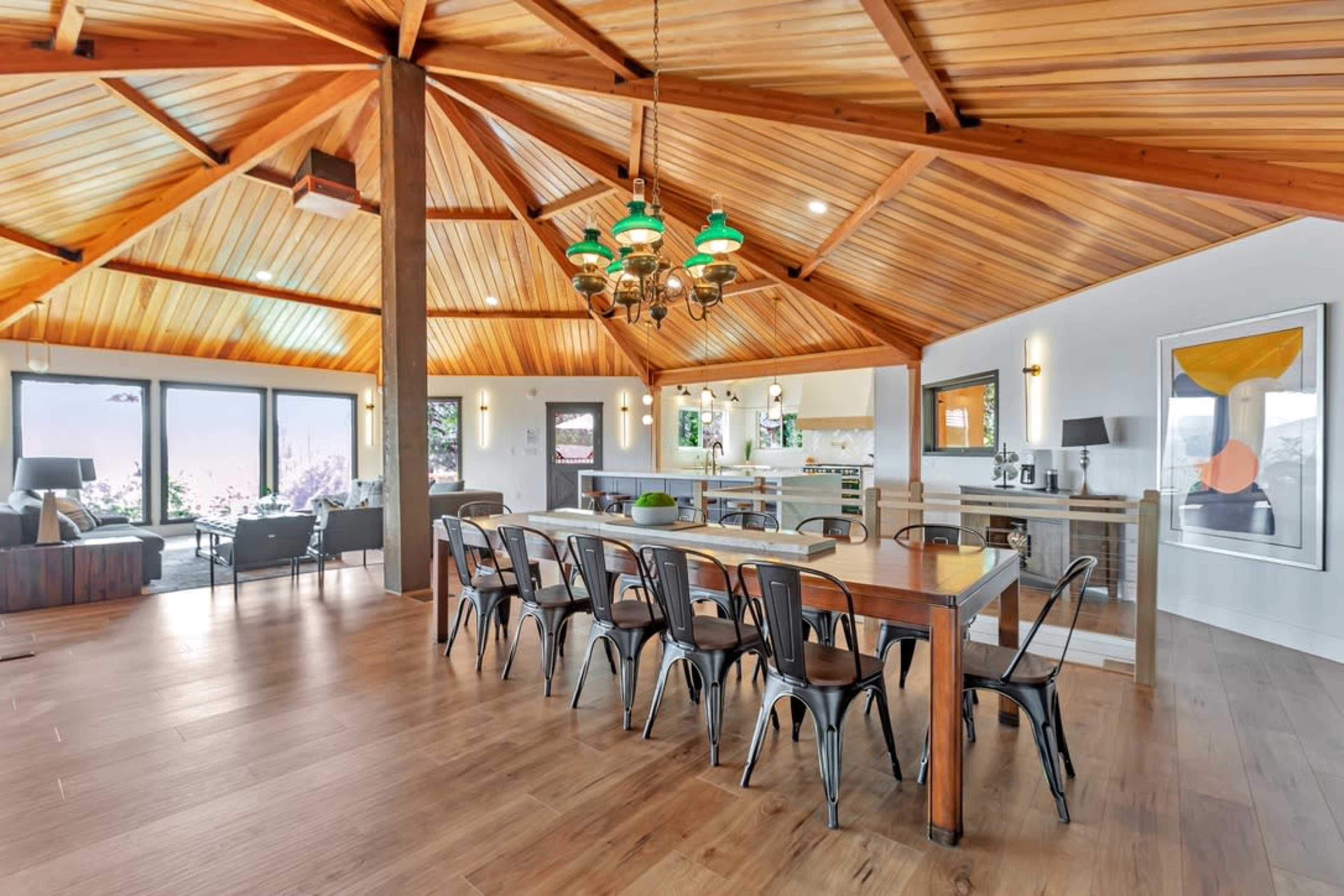 A spacious interior features a wooden ceiling with exposed beams, a long dining table with metal chairs, and an open layout connecting the kitchen and living area.