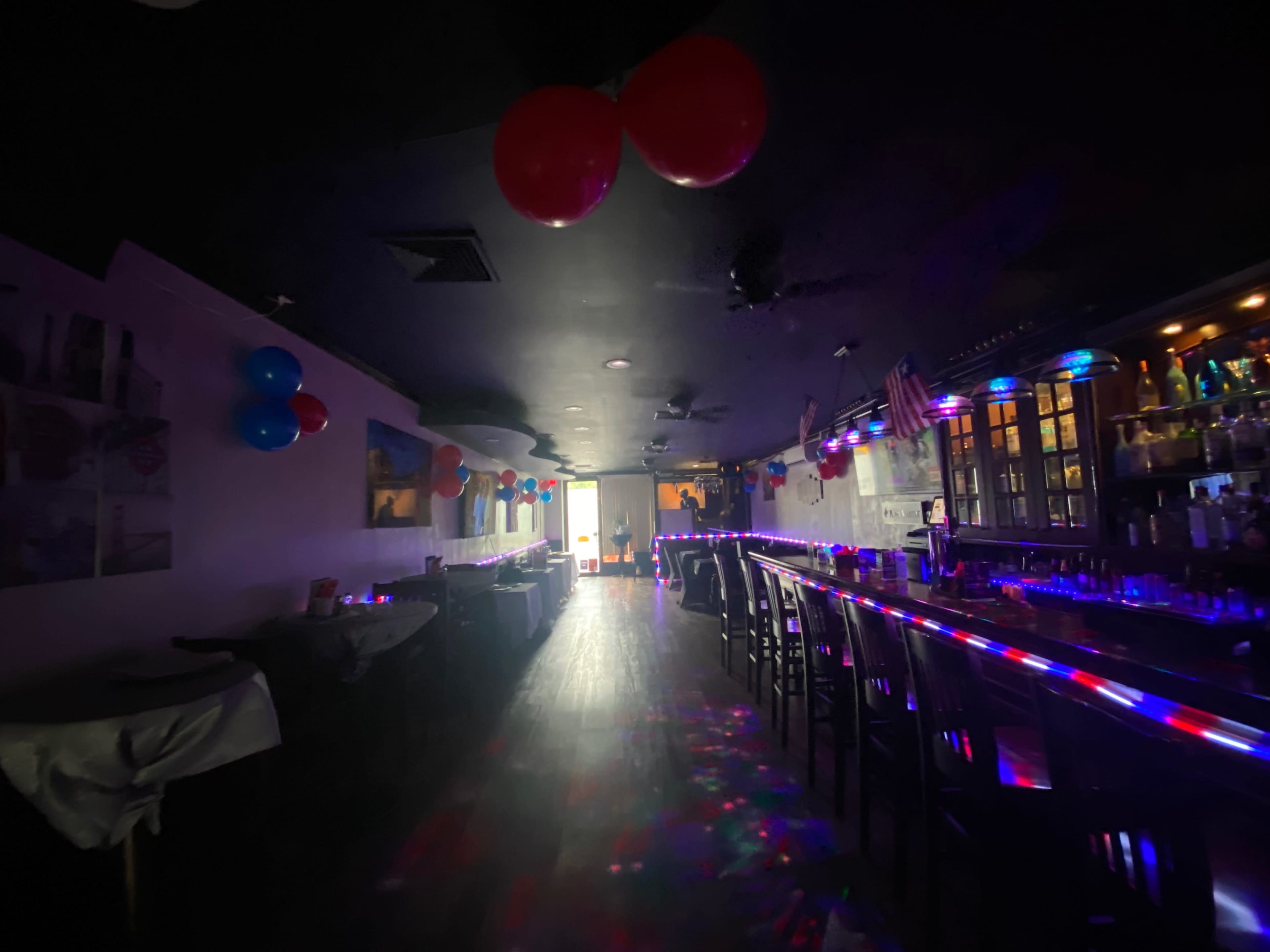 A dimly lit bar features a long counter with colorful lights and red and blue balloons decorating the ceiling.