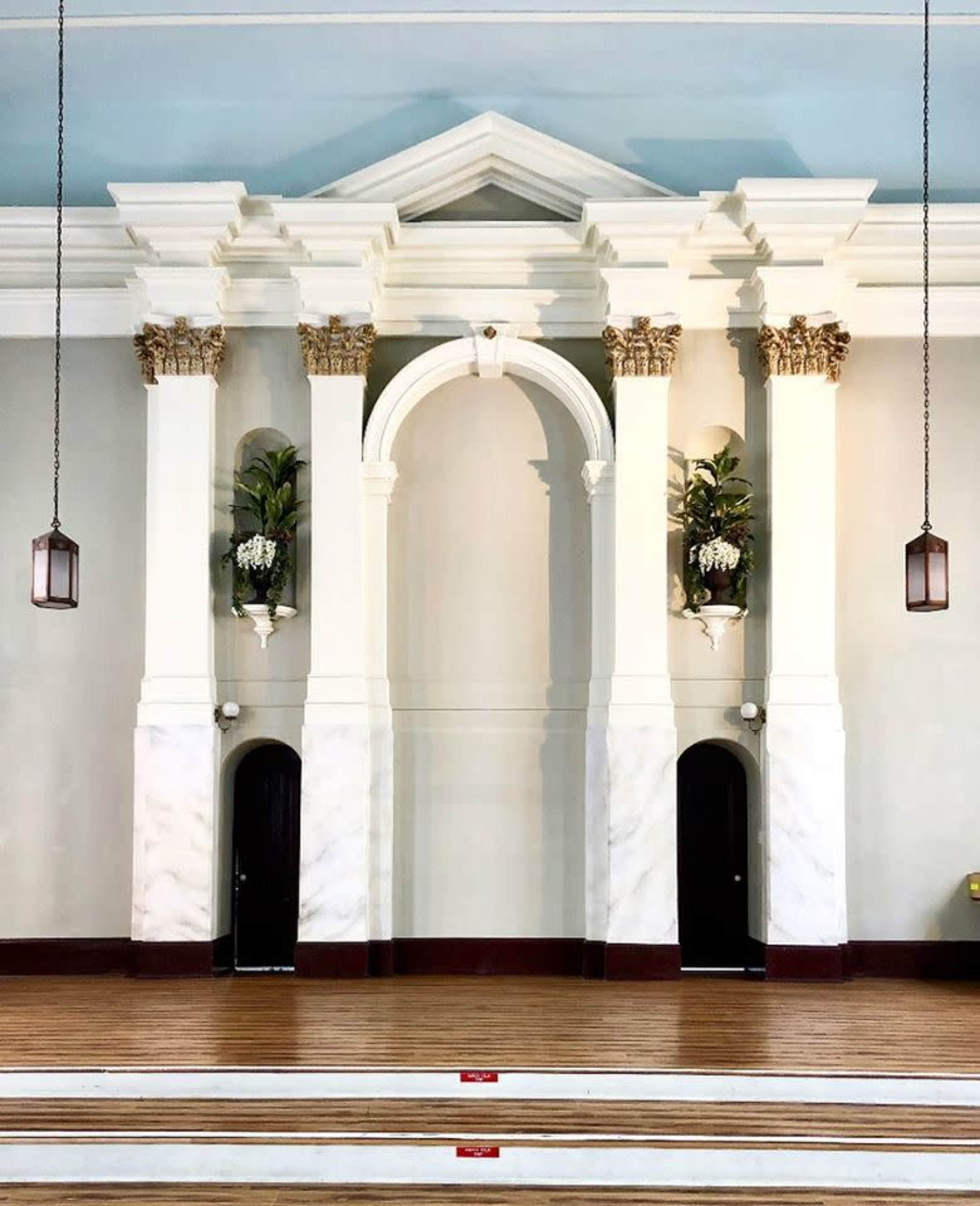 The image shows a large wall with an arched architectural feature flanked by two columns, each adorned with ornamental detailing and small plants, above a wooden floor with steps leading up to it.