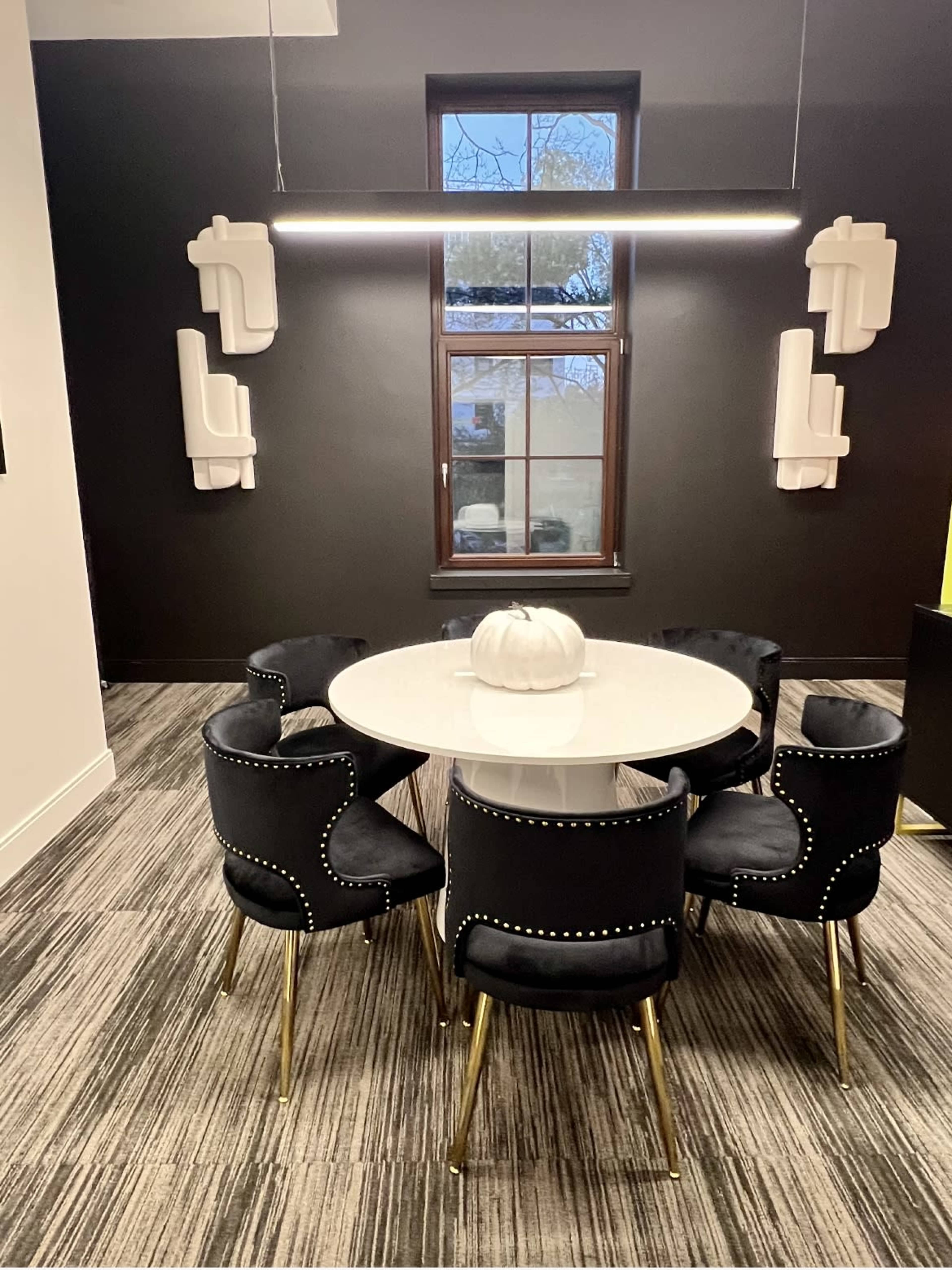 A round table with a white top is surrounded by six black chairs with gold legs in a room featuring dark walls and two decorative light fixtures.