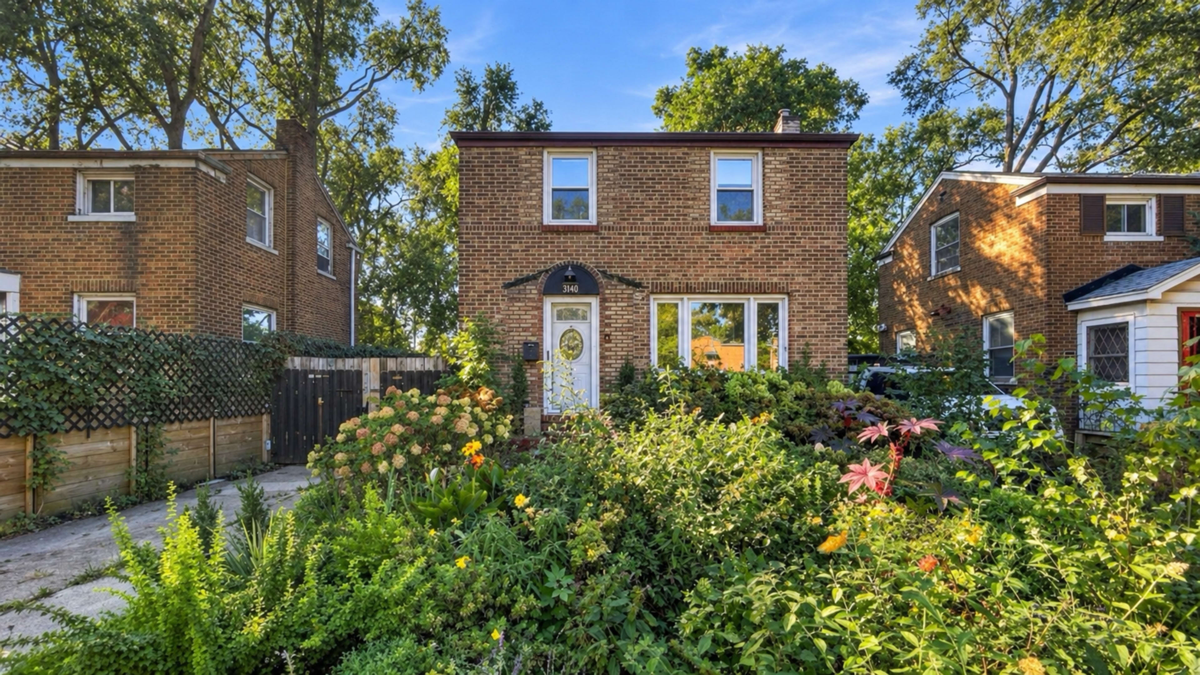 The image shows a two-story brick house surrounded by lush greenery and colorful flowers in the front yard.