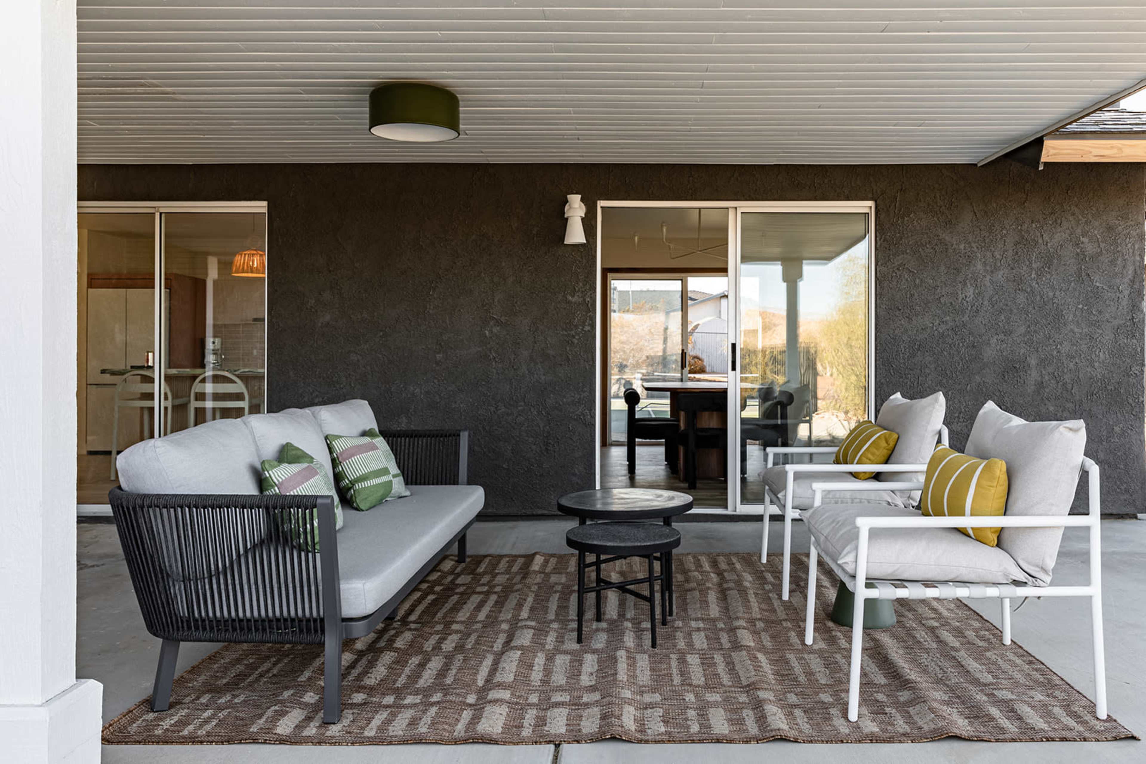 A covered patio features a modern seating arrangement with two chairs and a sofa, set around a small table on a geometric-patterned rug.