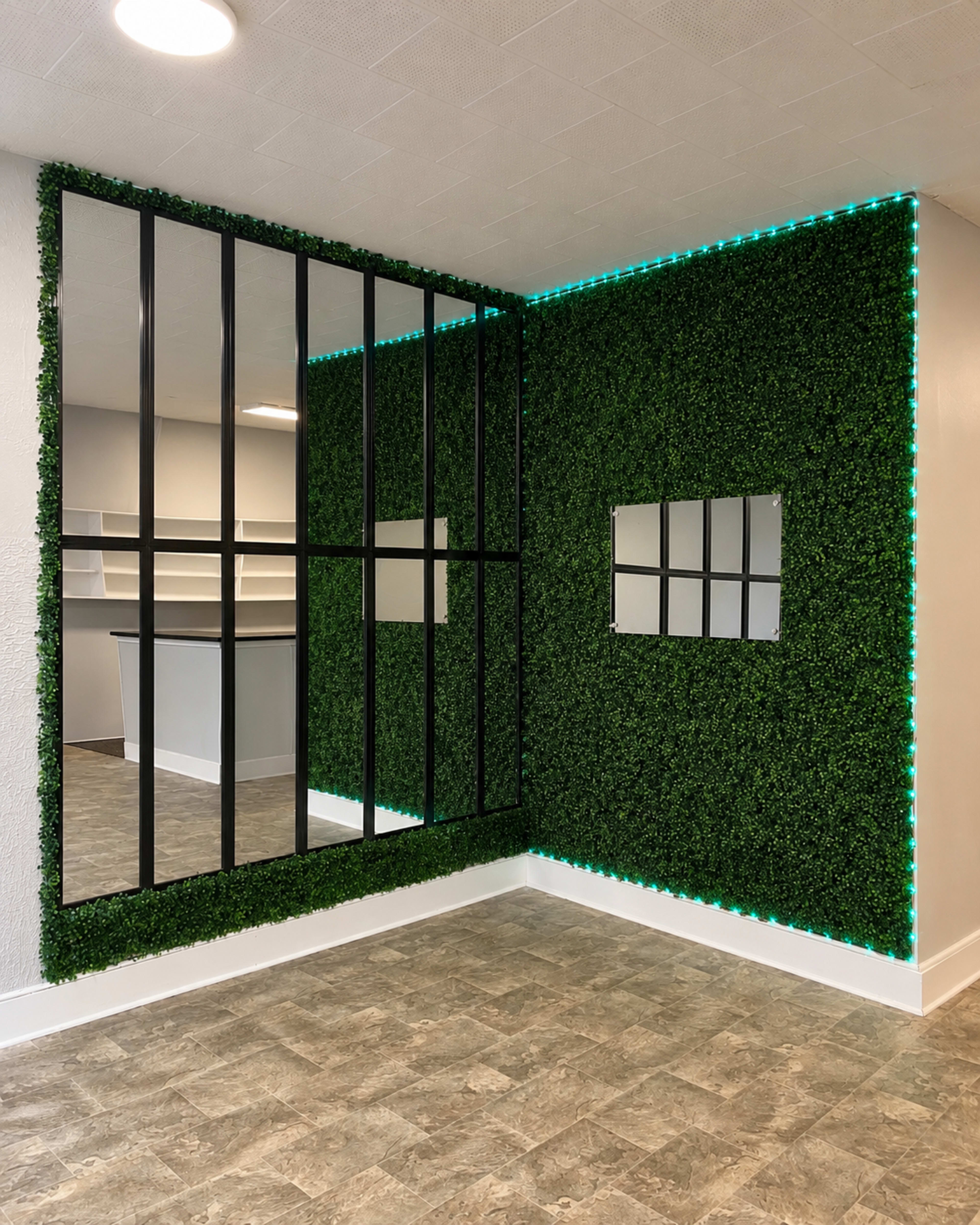 The image shows a corner of a room with a wall covered in artificial greenery, complemented by a grid-like black mirror and teal LED strip lighting.