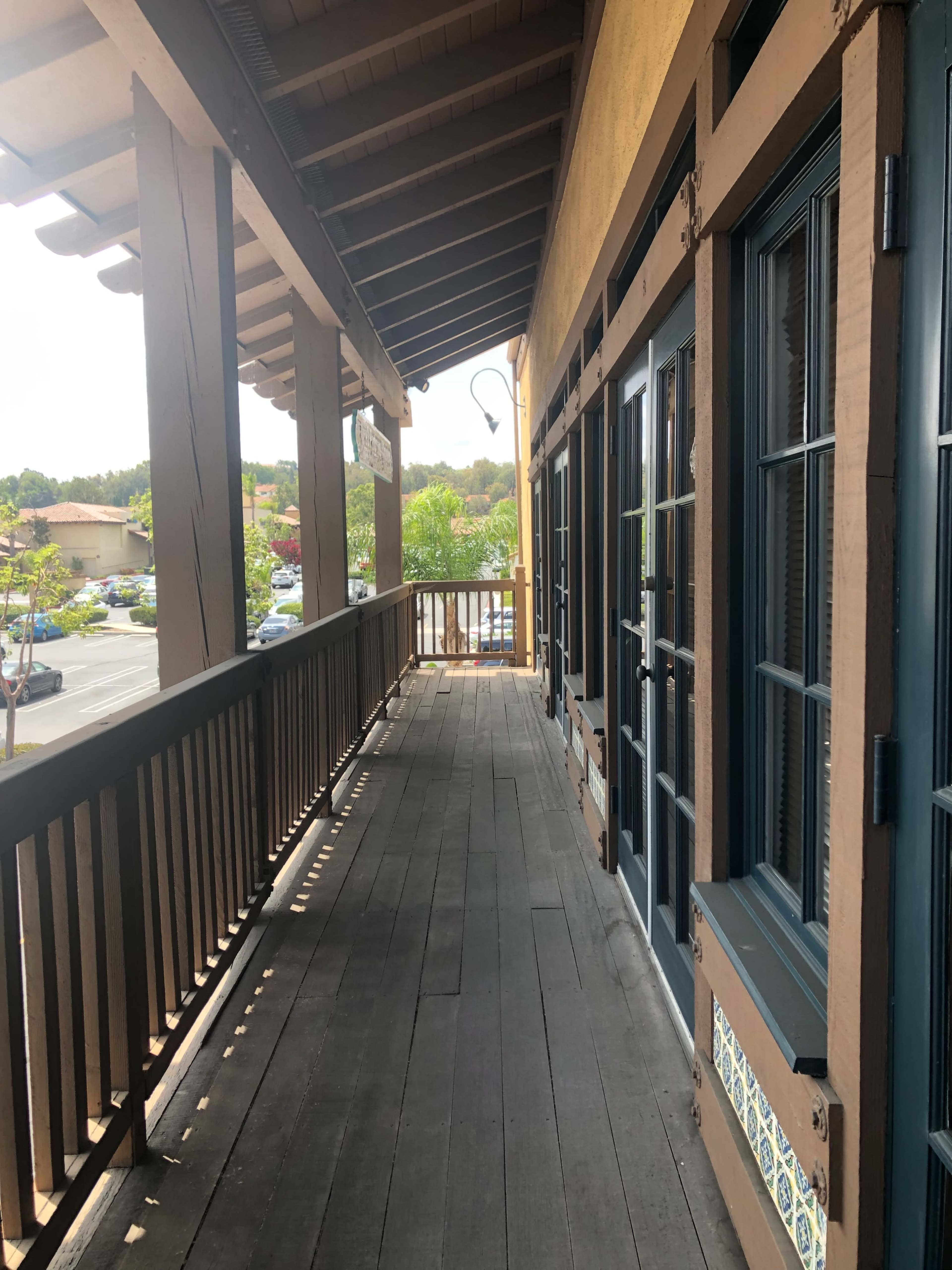 The image shows a long wooden balcony with doors on one side and a view of a parking lot in the background.