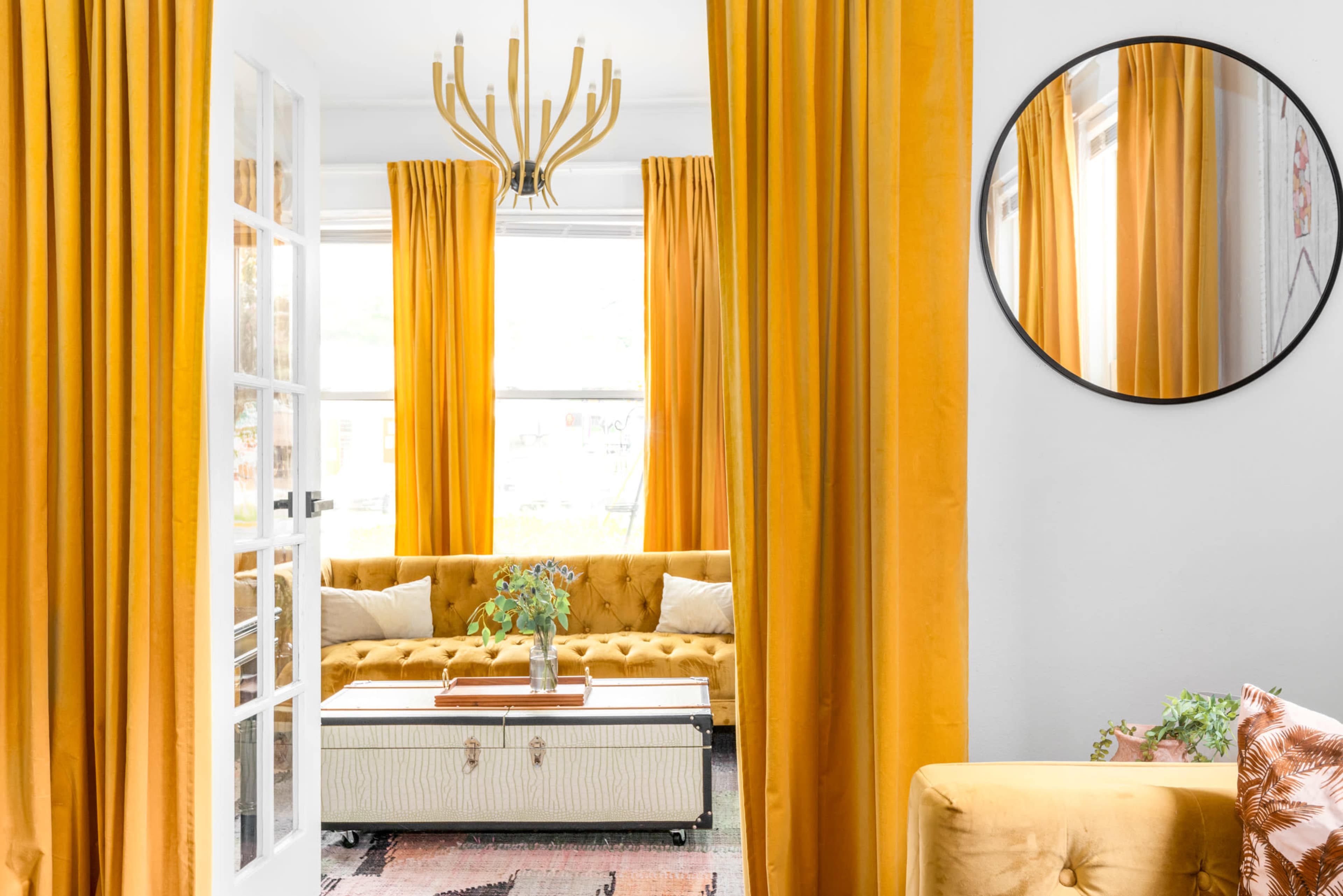 A bright living room features a yellow tufted sofa, long yellow curtains, and a round mirror, with a coffee table in the center.