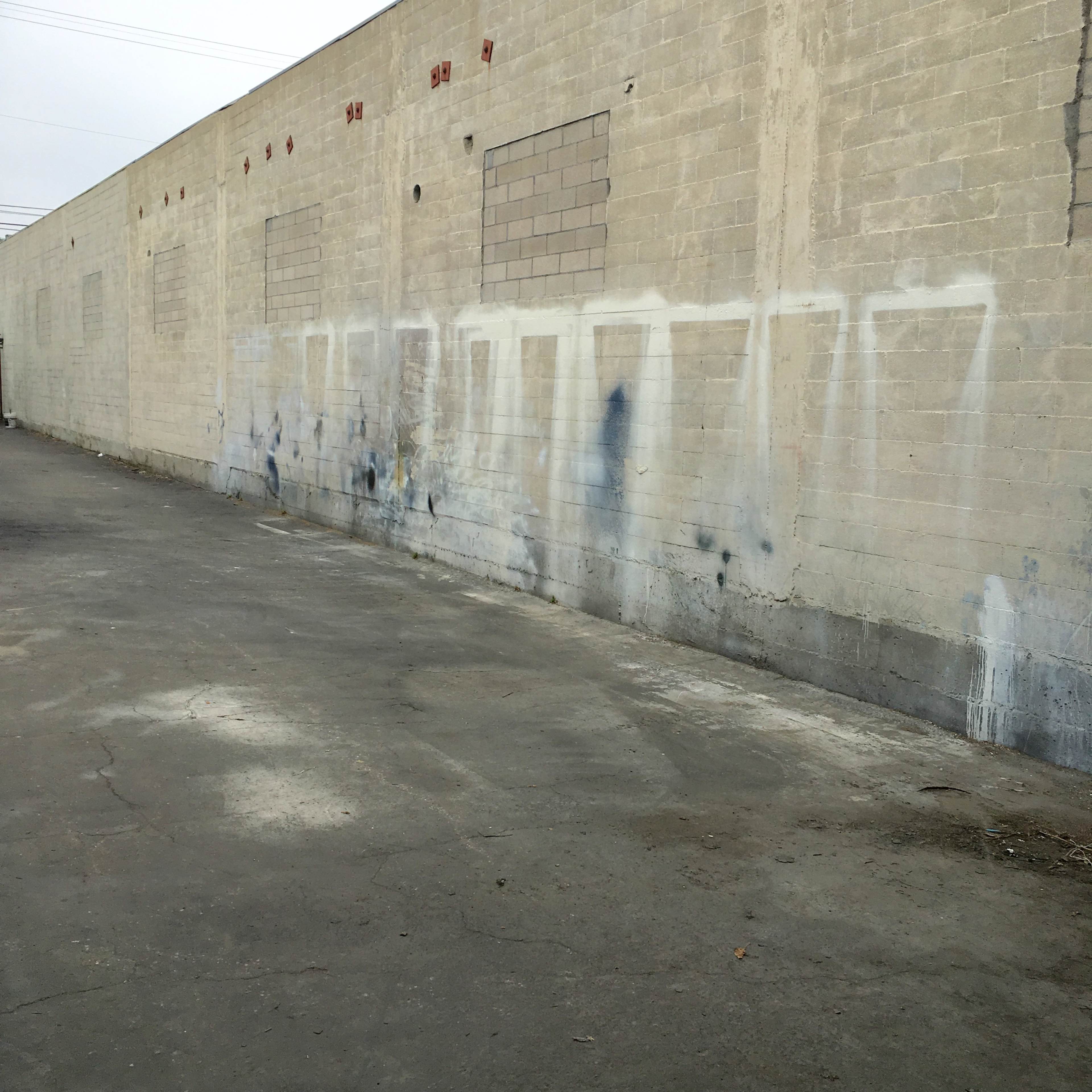 A weathered concrete wall with faded spray paint markings and a cracked surface, situated in an empty alleyway.