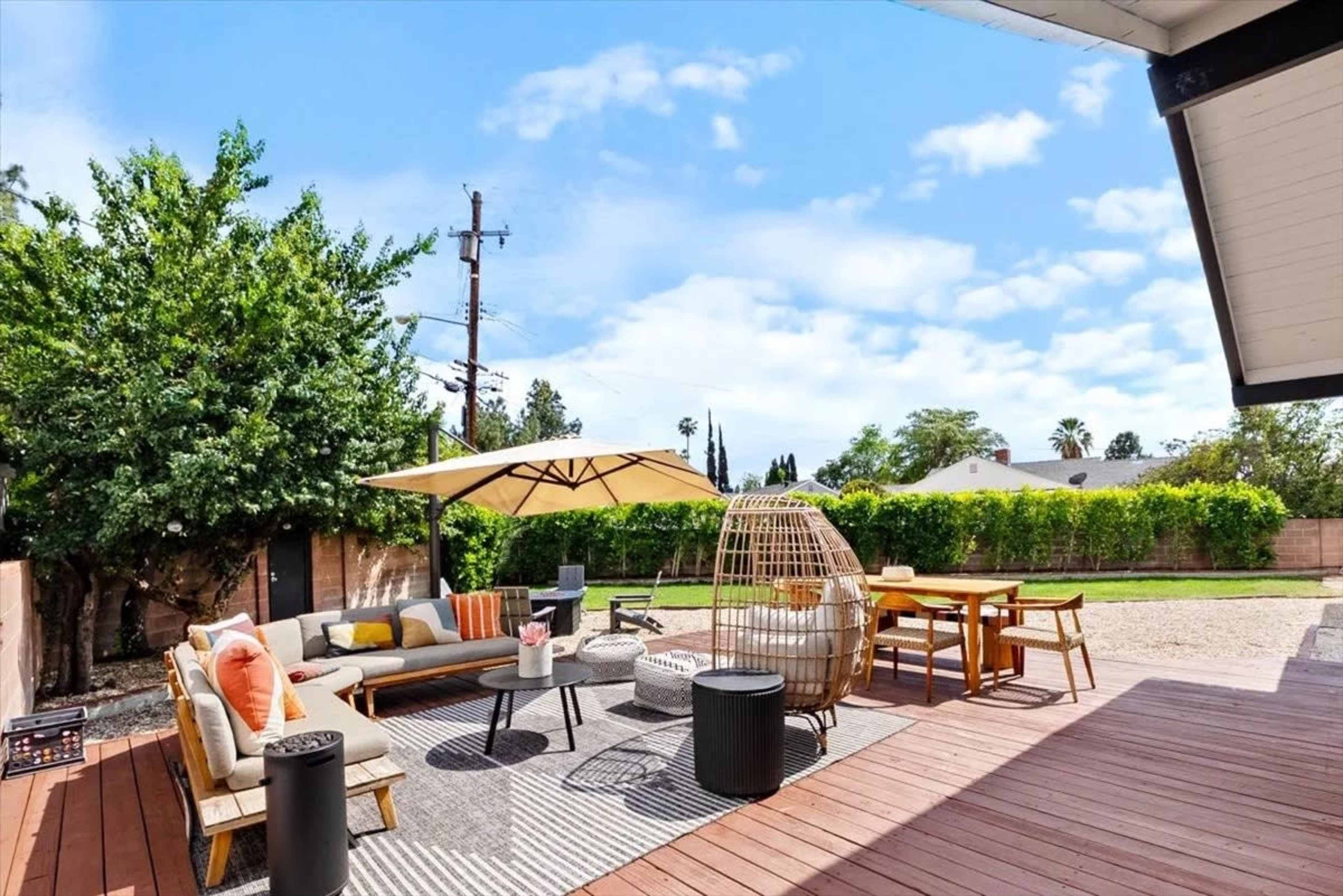 The image shows a spacious outdoor patio area with a couch, table, and chairs, surrounded by greenery and under a blue sky with a few clouds.