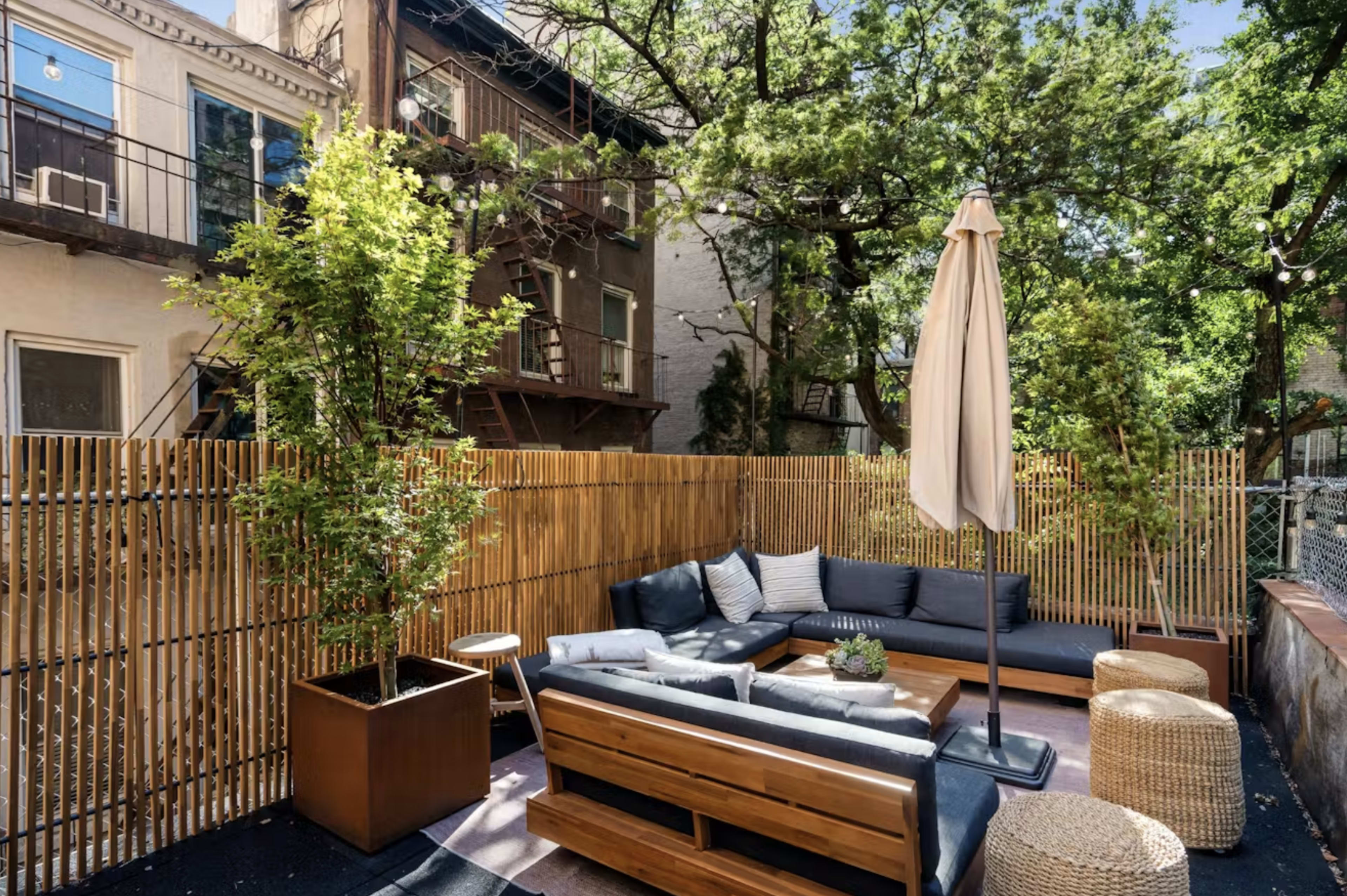 The image shows a cozy outdoor seating area with a sectional sofa, two wicker poufs, a potted plant, and a market umbrella, surrounded by wooden fencing and trees.