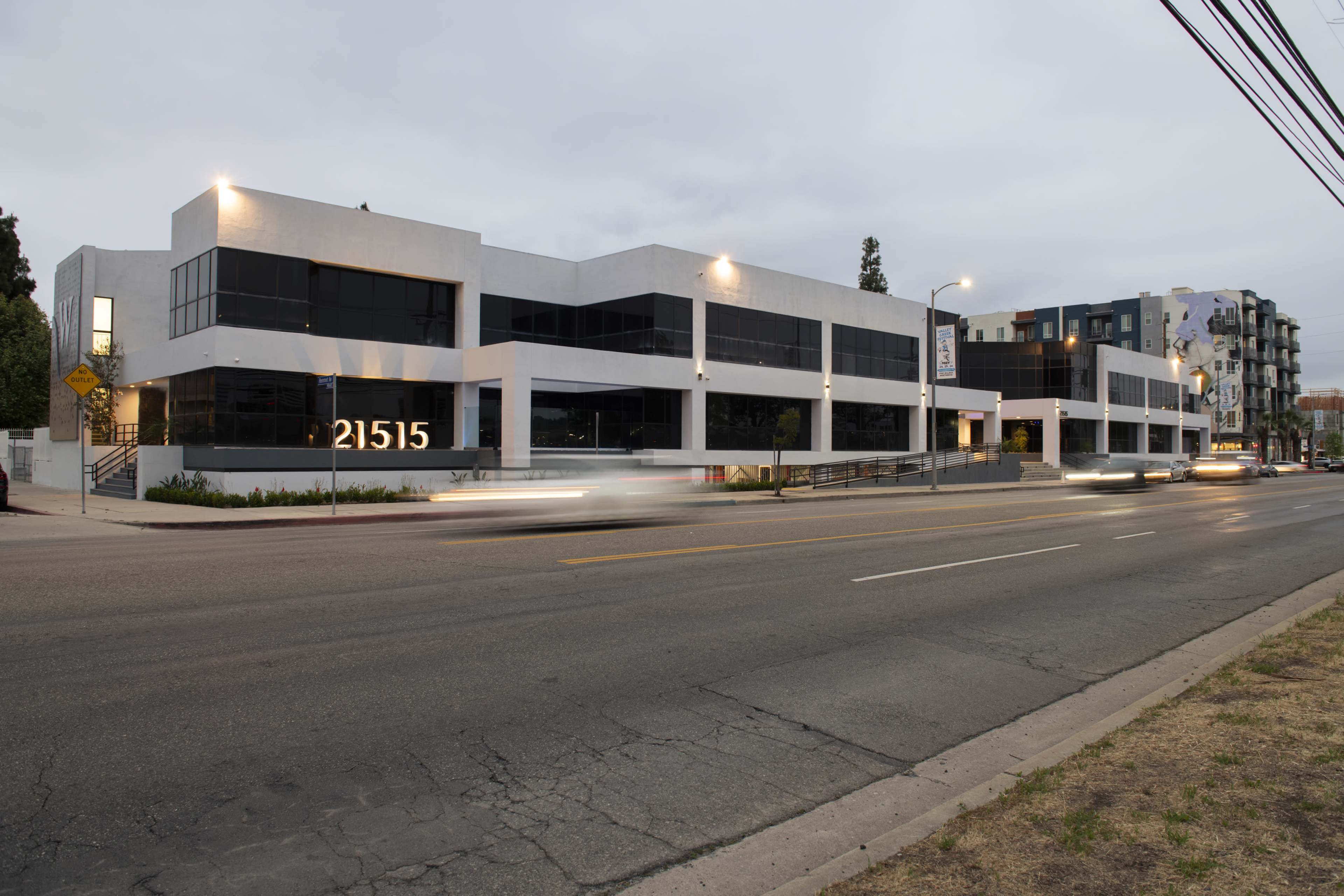 The image shows a modern, two-story commercial building with large windows, located on a street with moving traffic and a cloudy sky in the background.