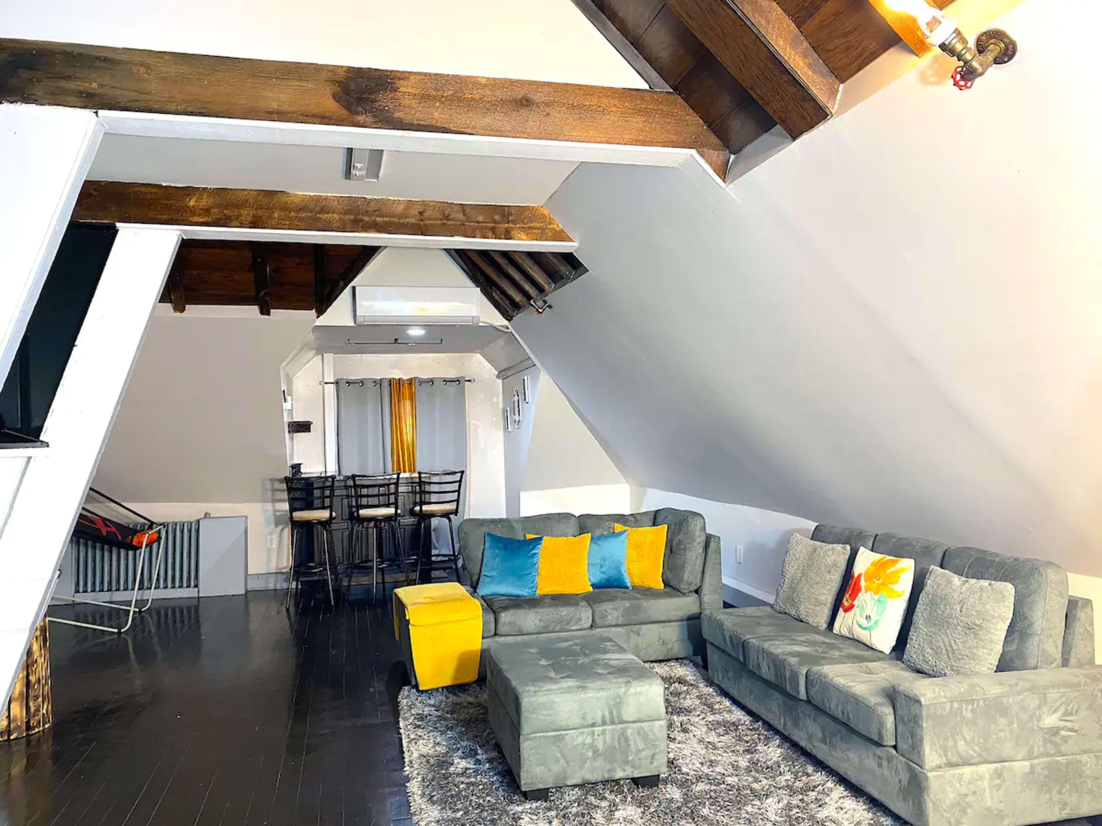 A cozy attic living space with sloped ceilings, a sectional sofa, a yellow ottoman, and a dining area with bar stools.