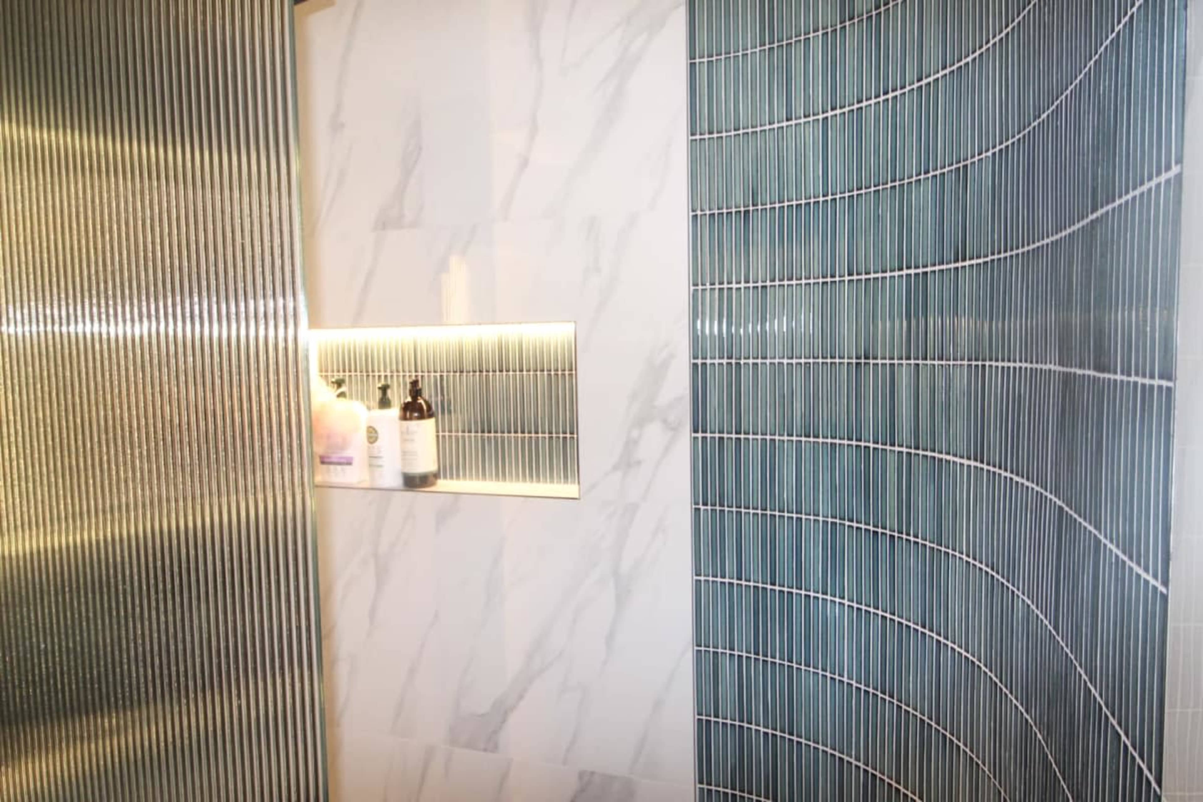 The image shows a modern shower space featuring two types of tile: a vertically striped design on one wall and a wave-patterned tile on the adjacent wall, with a recessed shelf displaying bottles.