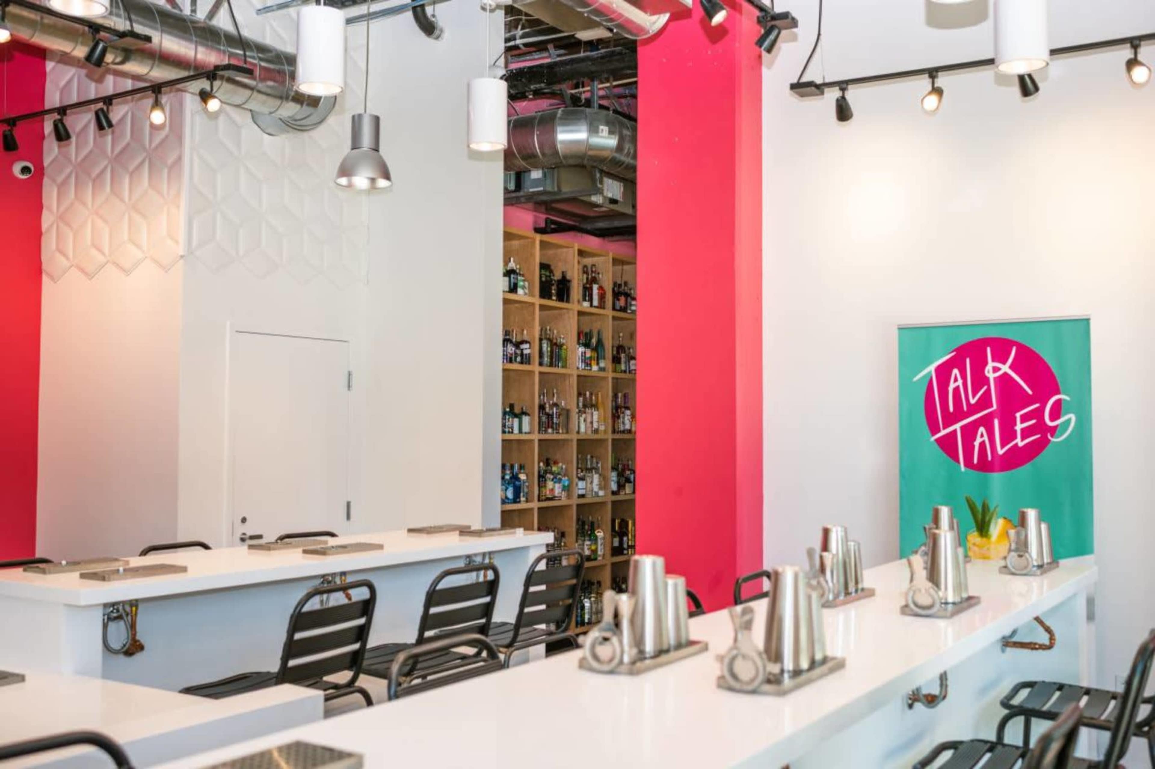 The image shows a modern bar area with a white counter, black chairs, and shelves lined with bottles against a bright pink wall.