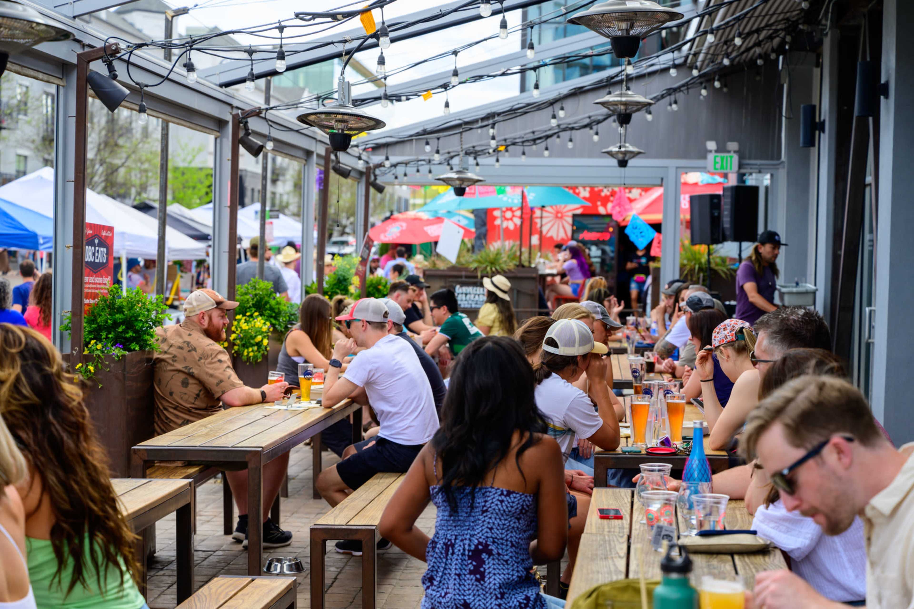 A bustling outdoor dining area filled with people sitting at wooden tables, enjoying food and drinks under hanging lights and heaters.