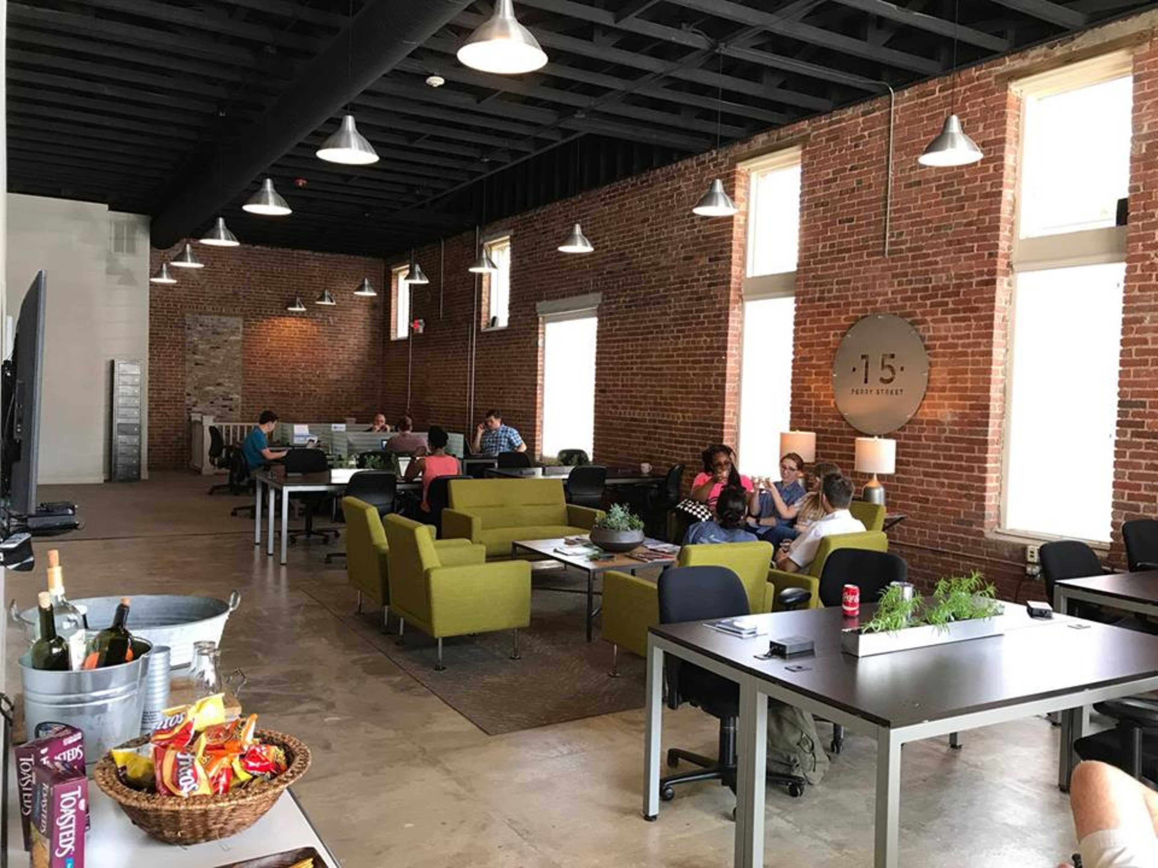 A spacious office with exposed brick walls is filled with people working at desks and sitting on green couches.