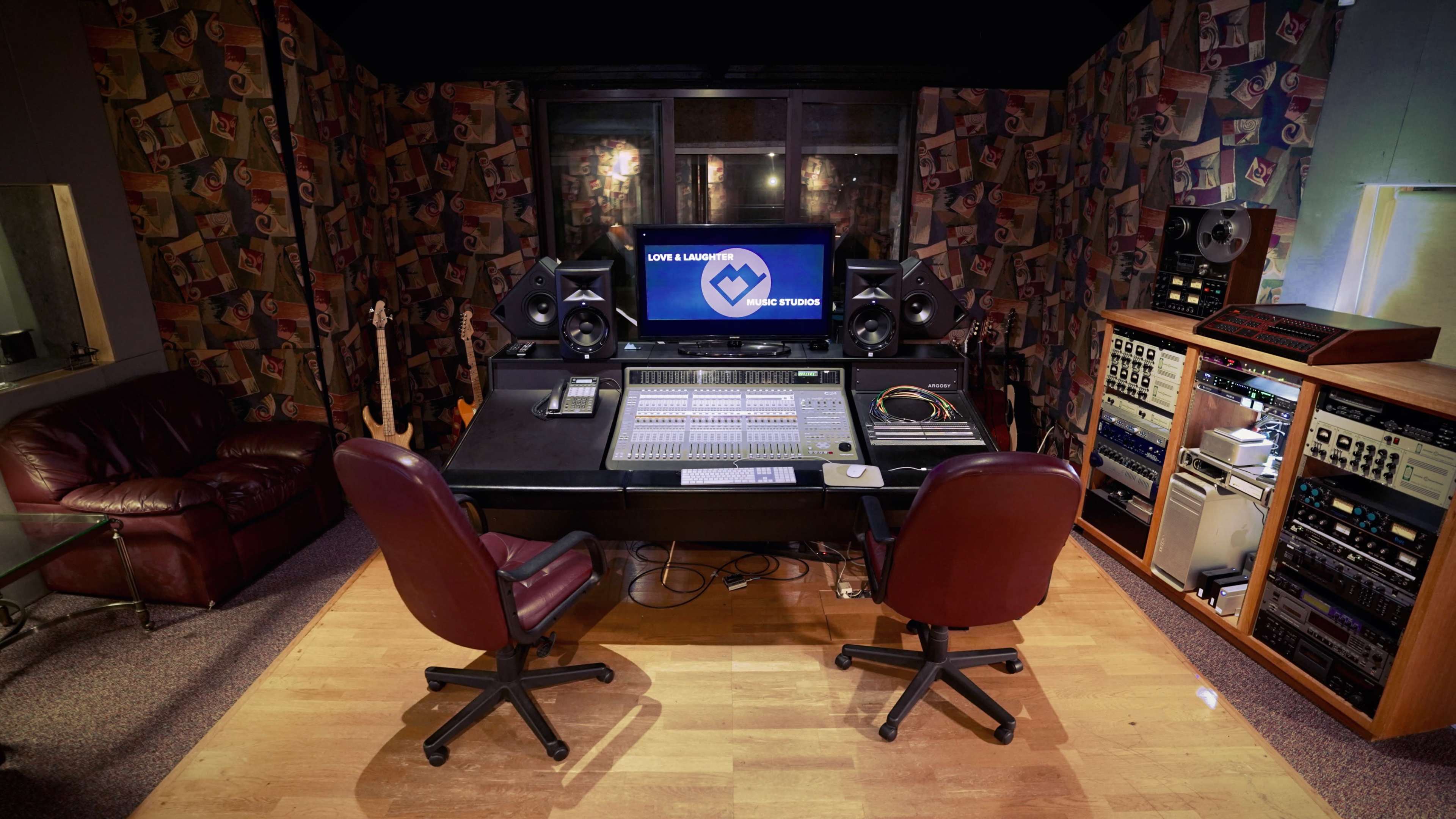 The image shows a music recording studio with a large mixing console, computer screen, and two red chairs facing the console, surrounded by colorful soundproofing panels.