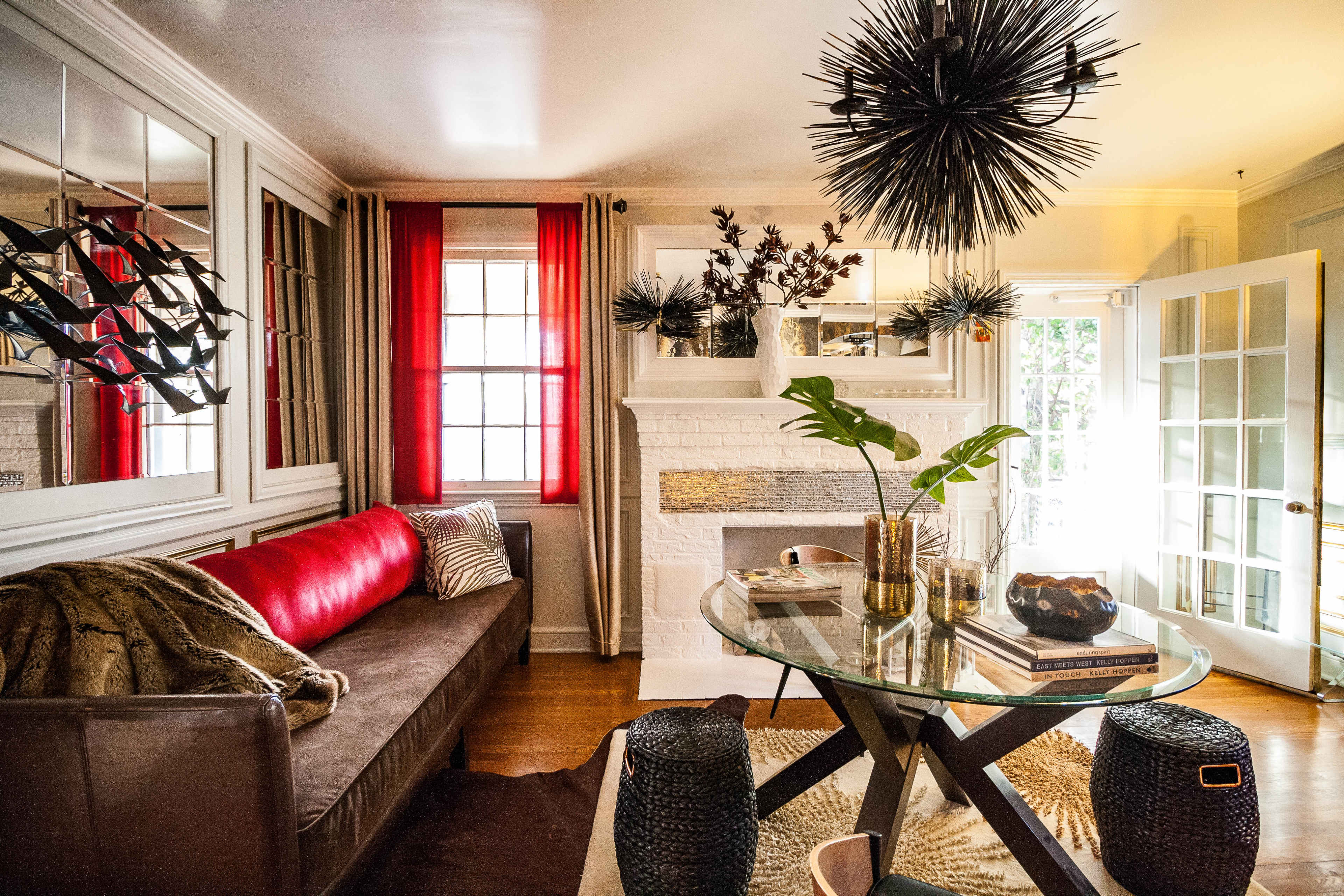 A modern living room featuring a brown sofa with red accents, a glass table with decorative items, and large windows with sheer curtains.