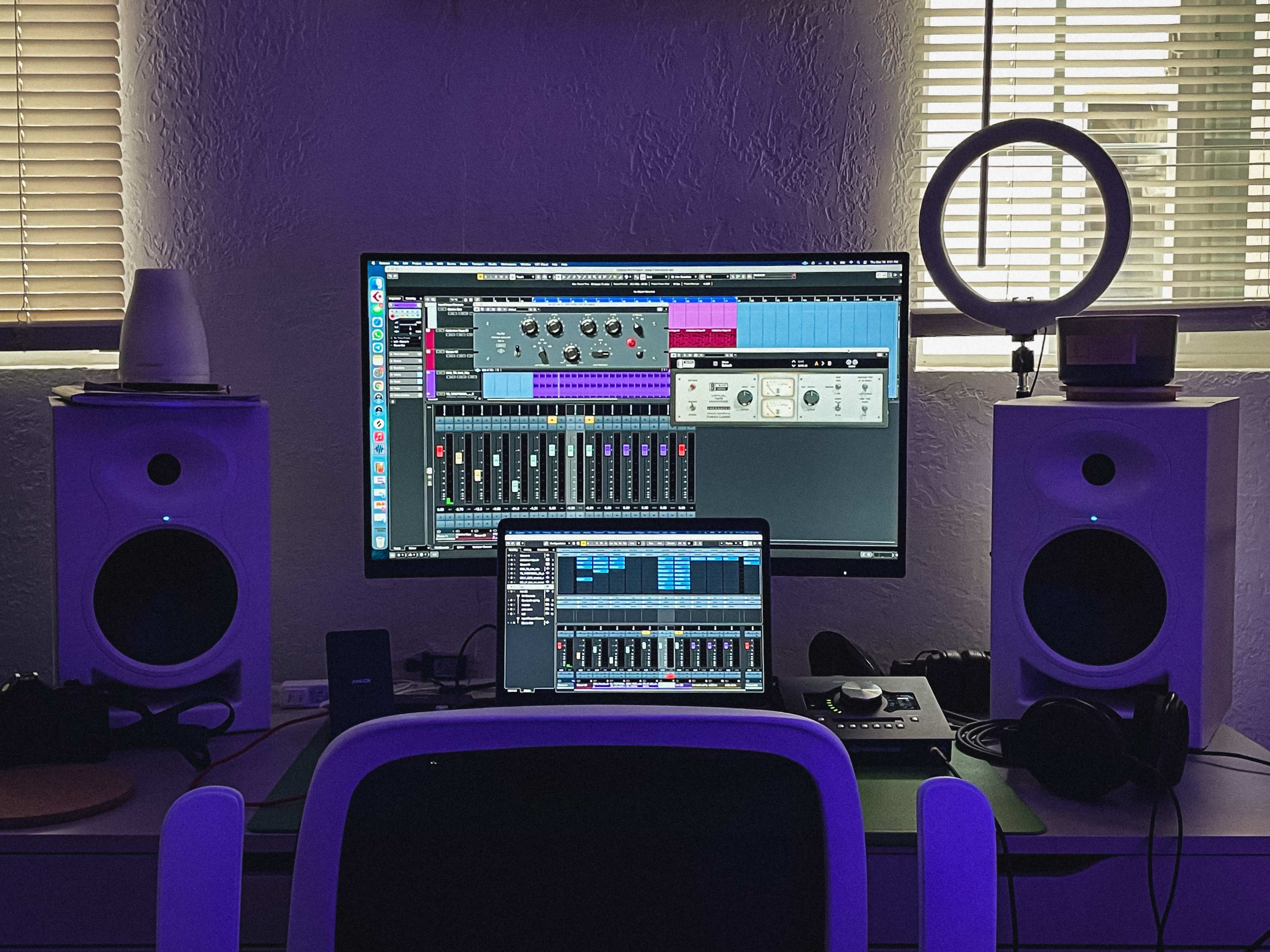 A home studio setup features a large monitor displaying audio editing software, a laptop, and two studio monitors, with a ring light illuminating the workspace.