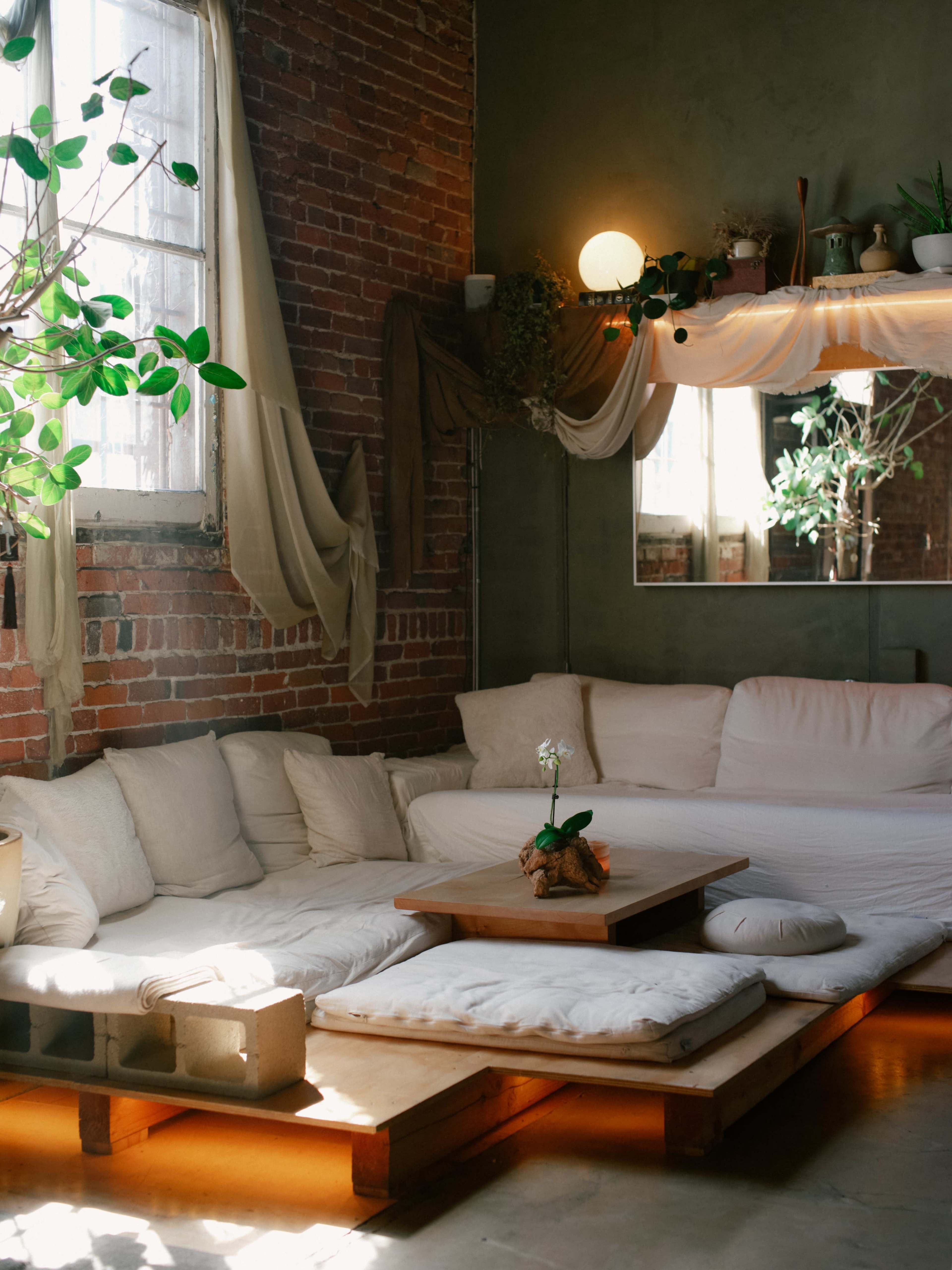 The image shows a cozy, minimalist living space with a large cushioned seating area, a wooden table, and several plants near a window.