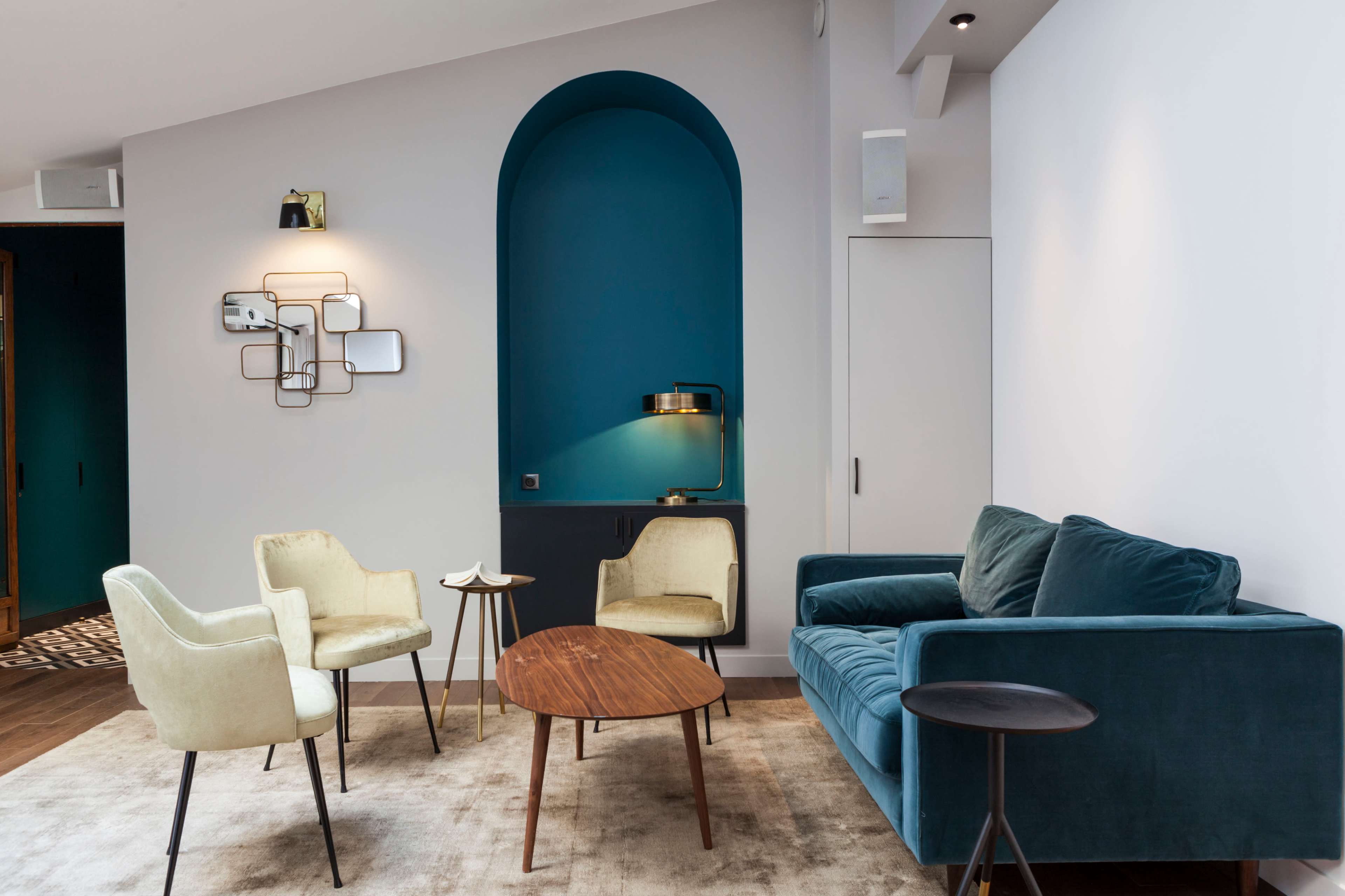 The image shows a modern living space with a teal accent wall, a blue sofa, two cream-colored chairs, a round wooden coffee table, and a small side table.