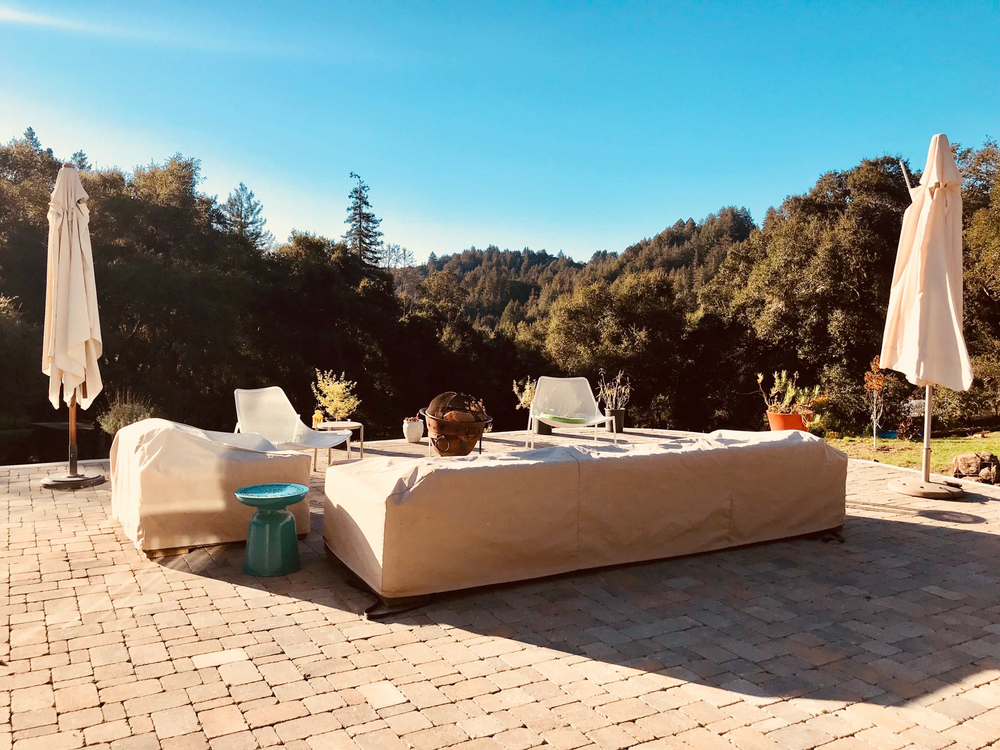 The image shows a covered outdoor seating area with a table and chairs, surrounded by lush greenery and blue skies.