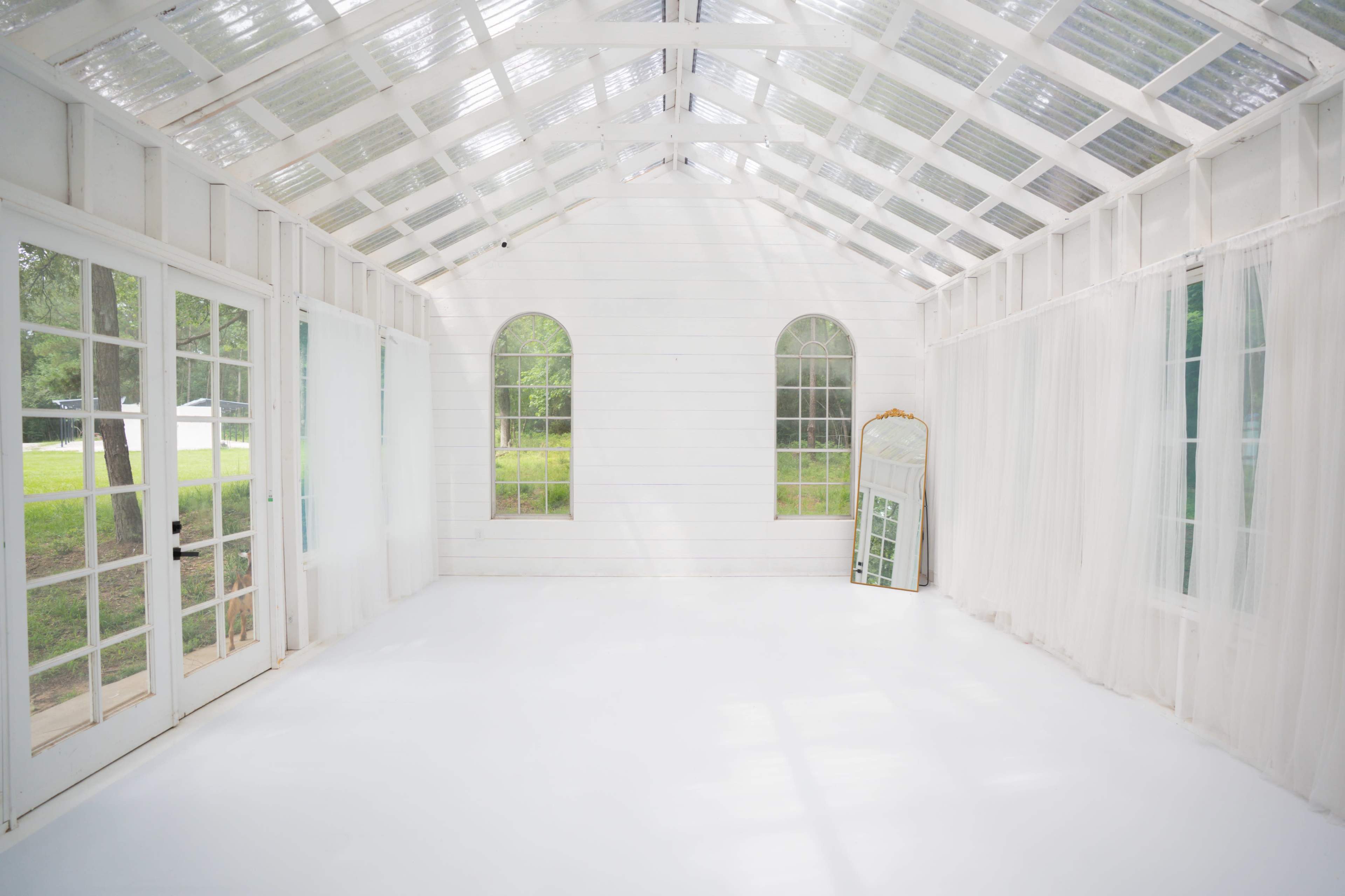 The image shows a spacious, white interior with large windows, sheer curtains, and a large mirror propped against the wall.
