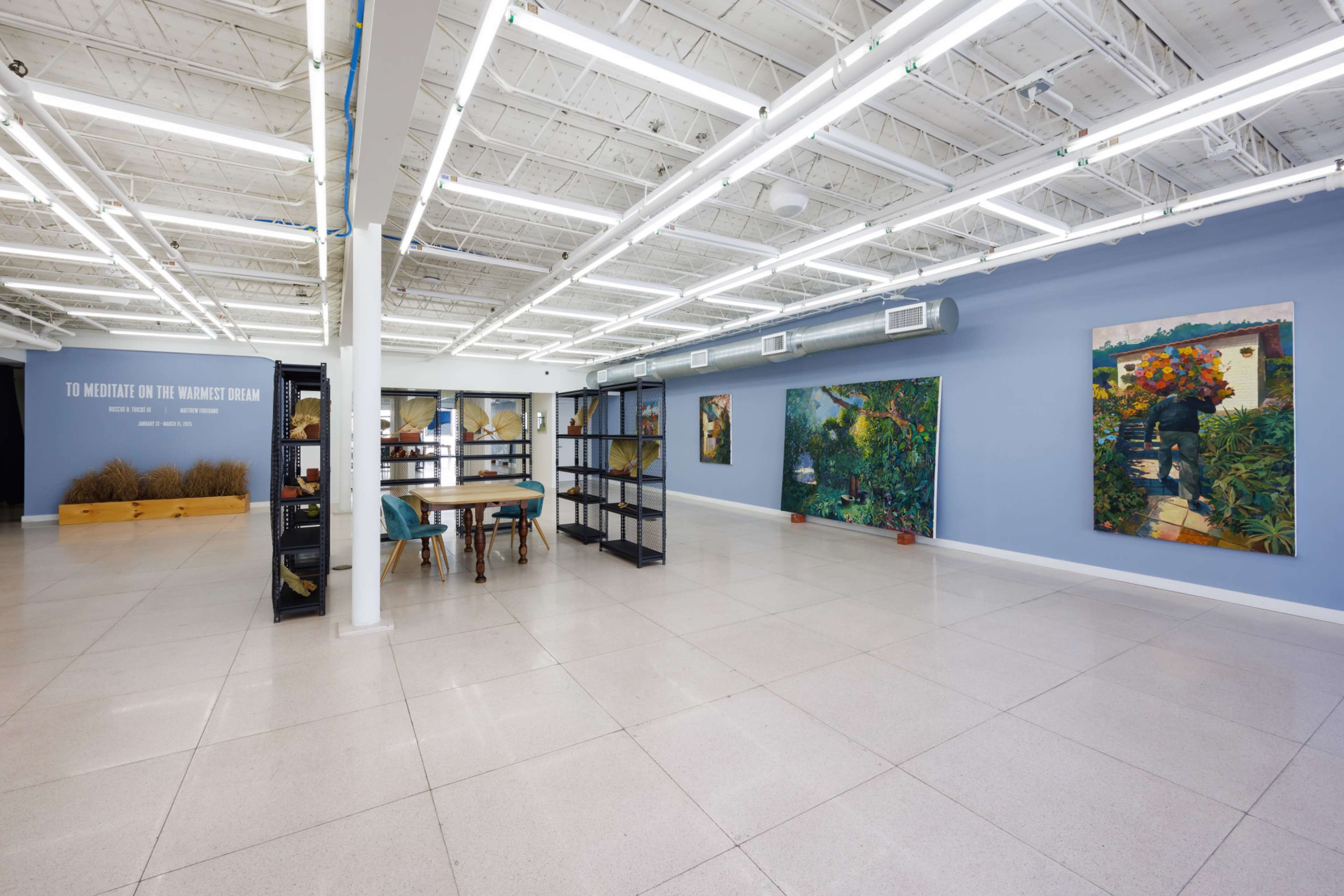 The image shows a spacious, well-lit art gallery featuring two large paintings on the walls and a central table with chairs.