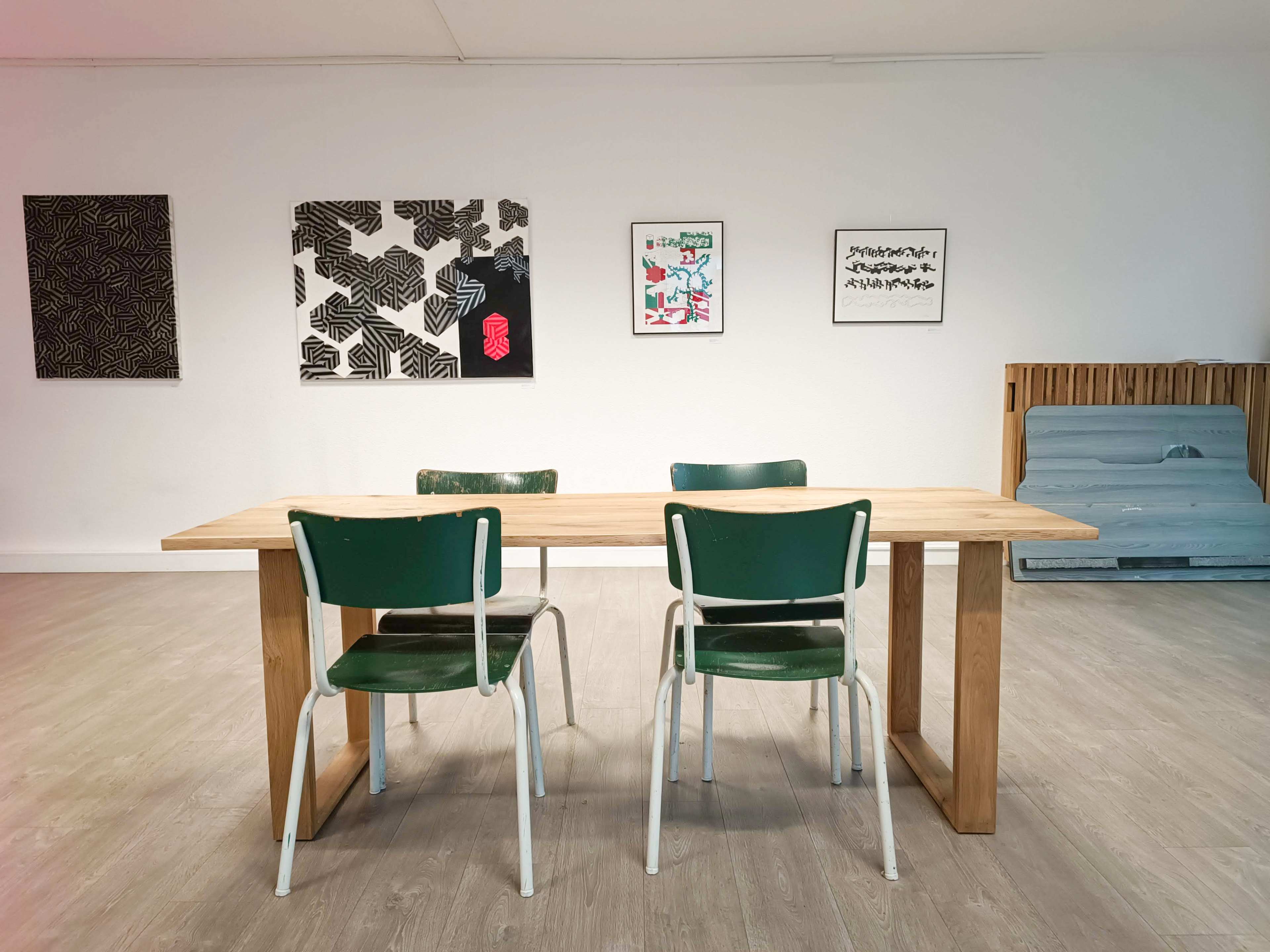 A wooden table with two green chairs sits in an art-filled room featuring various framed artworks on the walls.