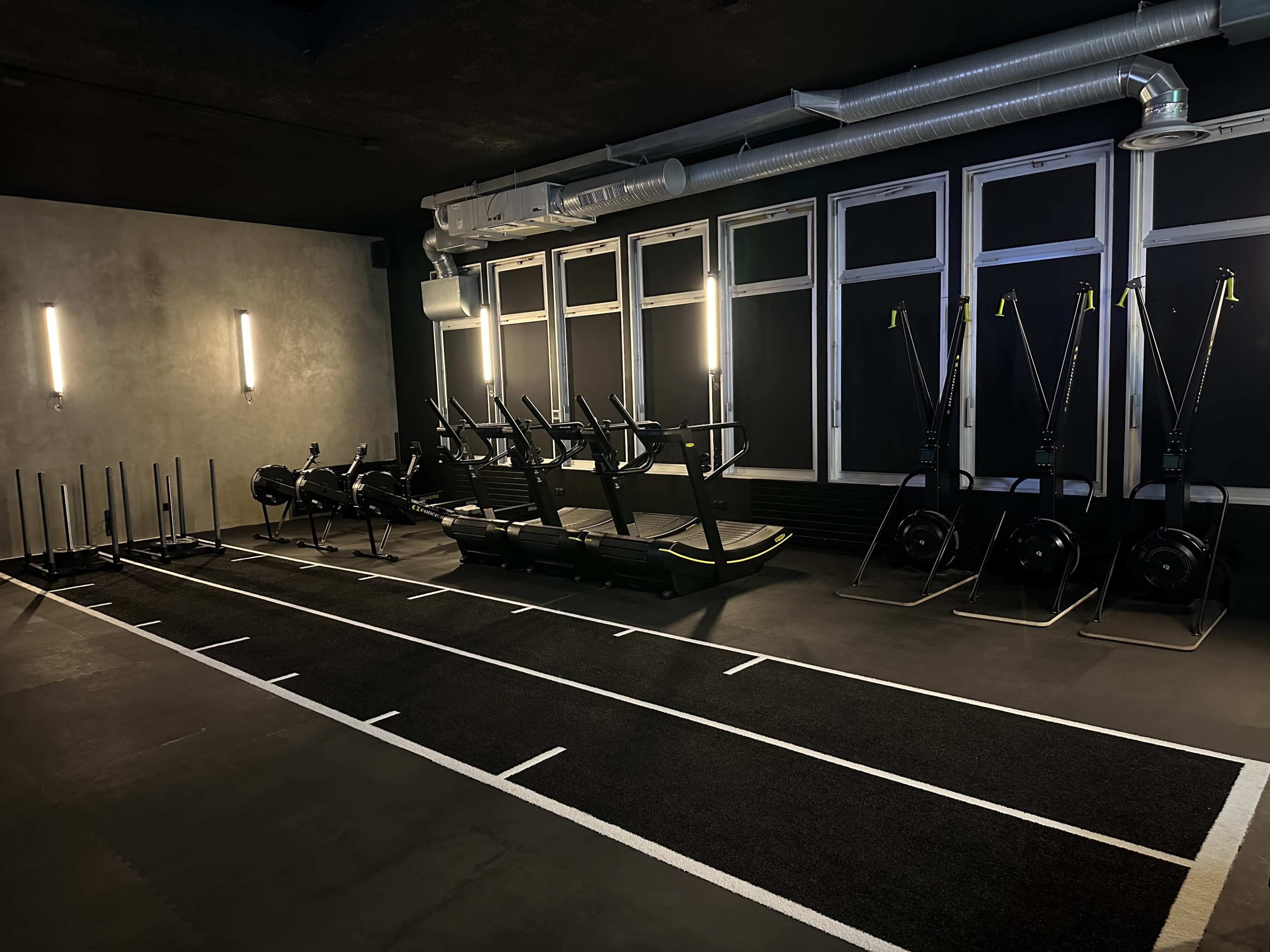 The image shows a modern gym interior featuring exercise equipment such as rowing machines, treadmills, and sleds arranged neatly in a well-lit space.