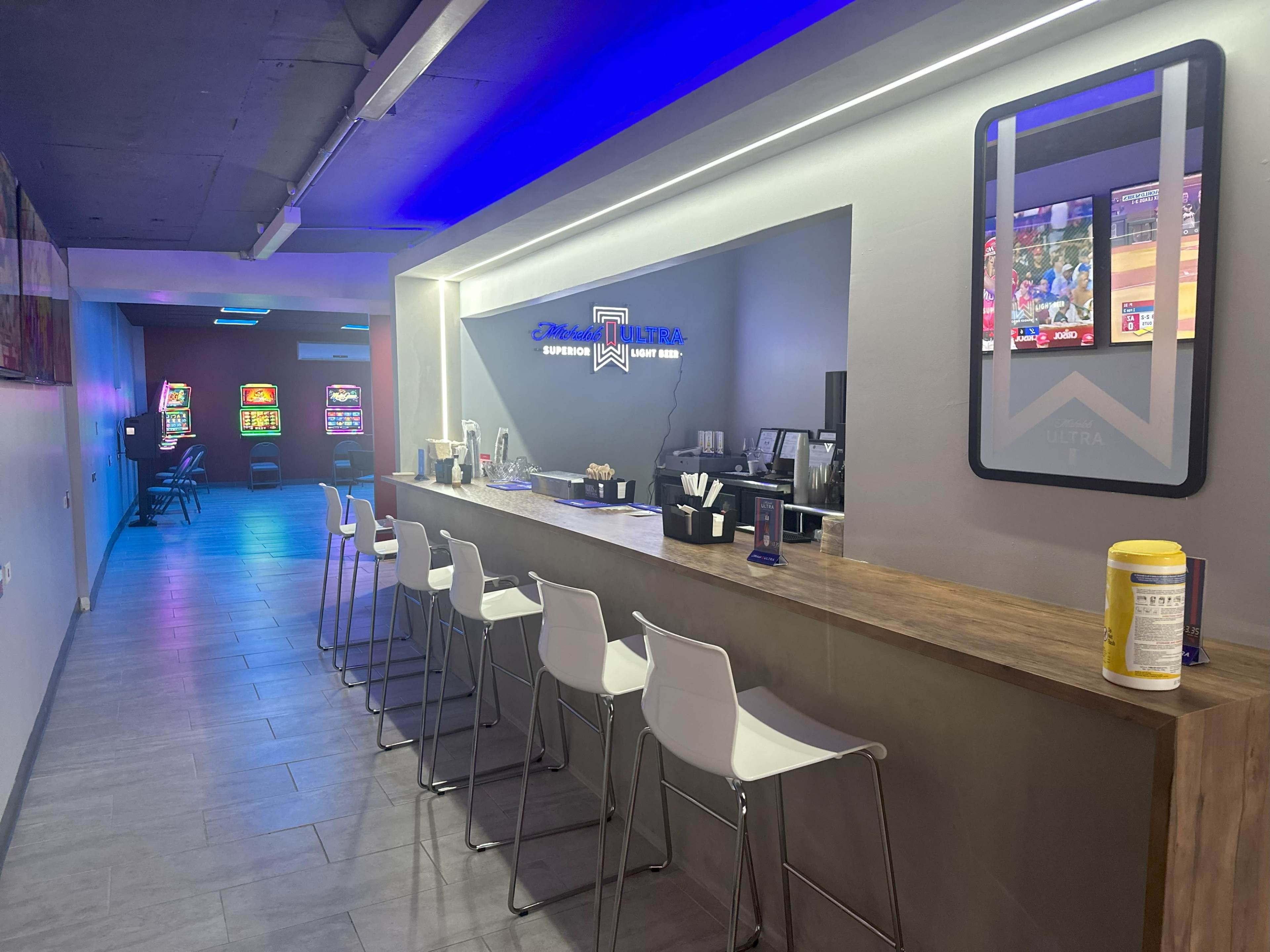 The image shows a modern lounge area with a bar featuring high stools, illuminated by soft lighting, and a hallway leading to a gaming room with arcade machines.
