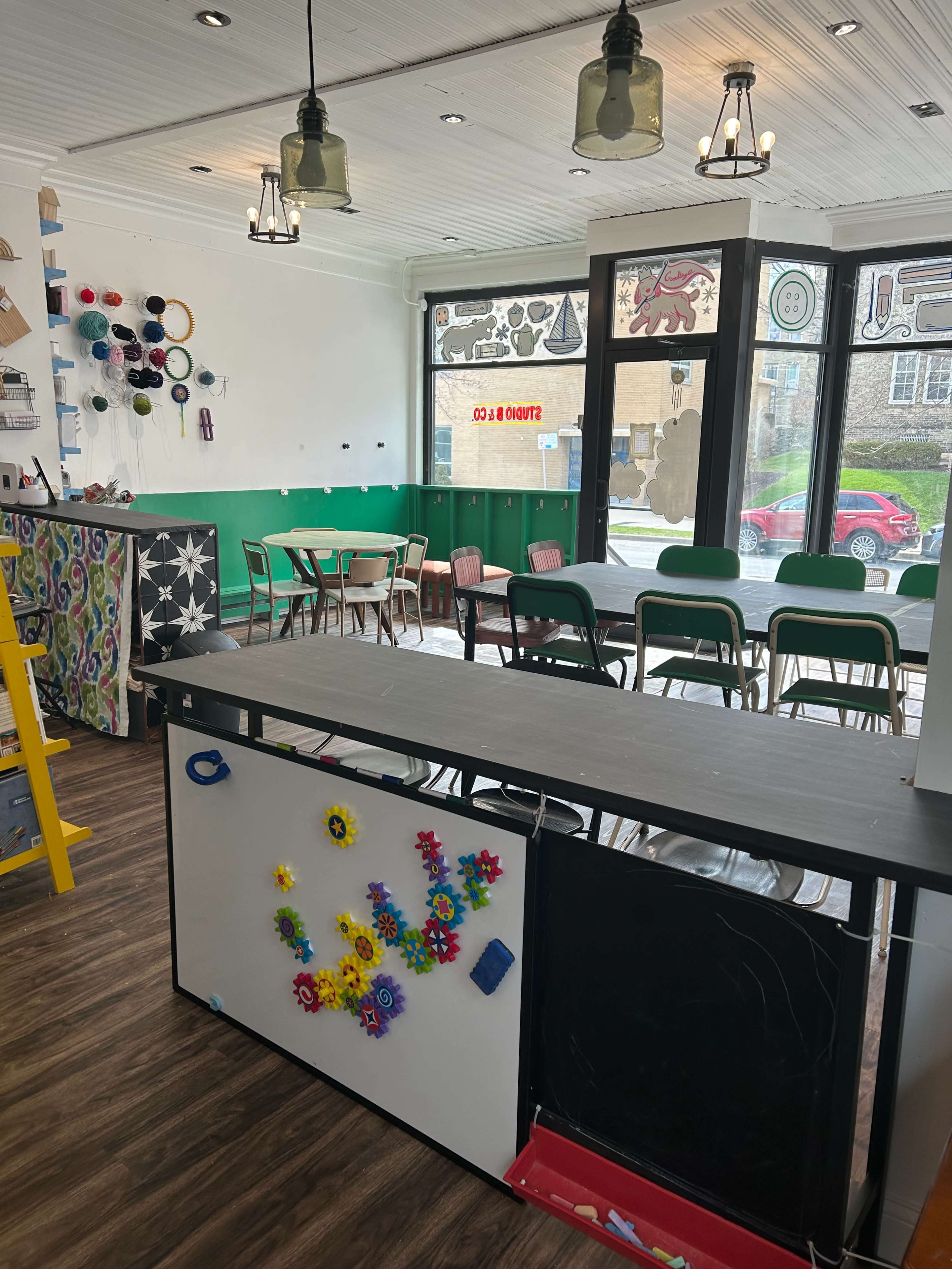 The image shows an interior of a colorful café or community space with a seating area, a counter adorned with decorative items, and large windows allowing natural light in.