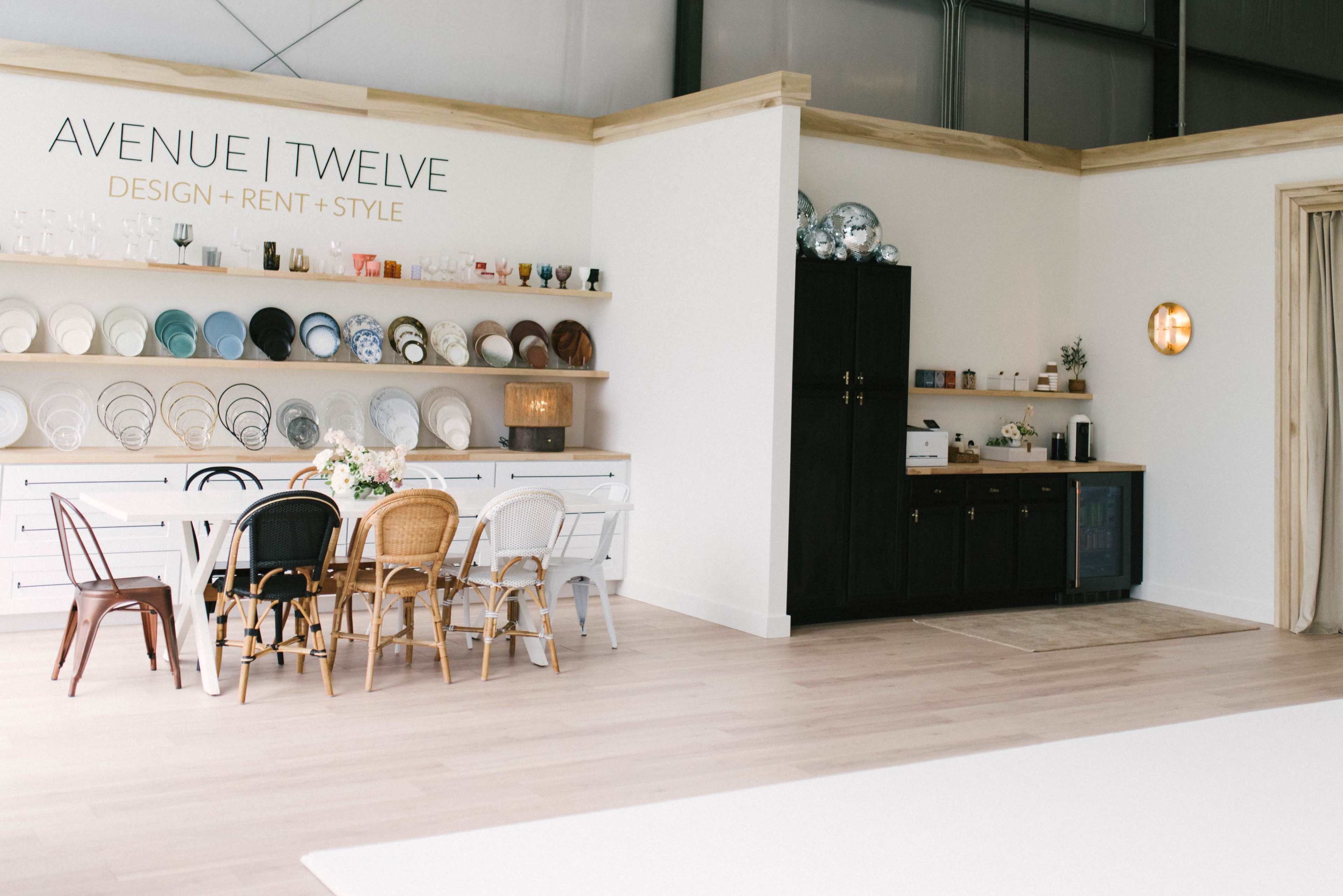 A bright interior space featuring a decorated wall with various plates, a dining table surrounded by chairs, and a kitchenette area.