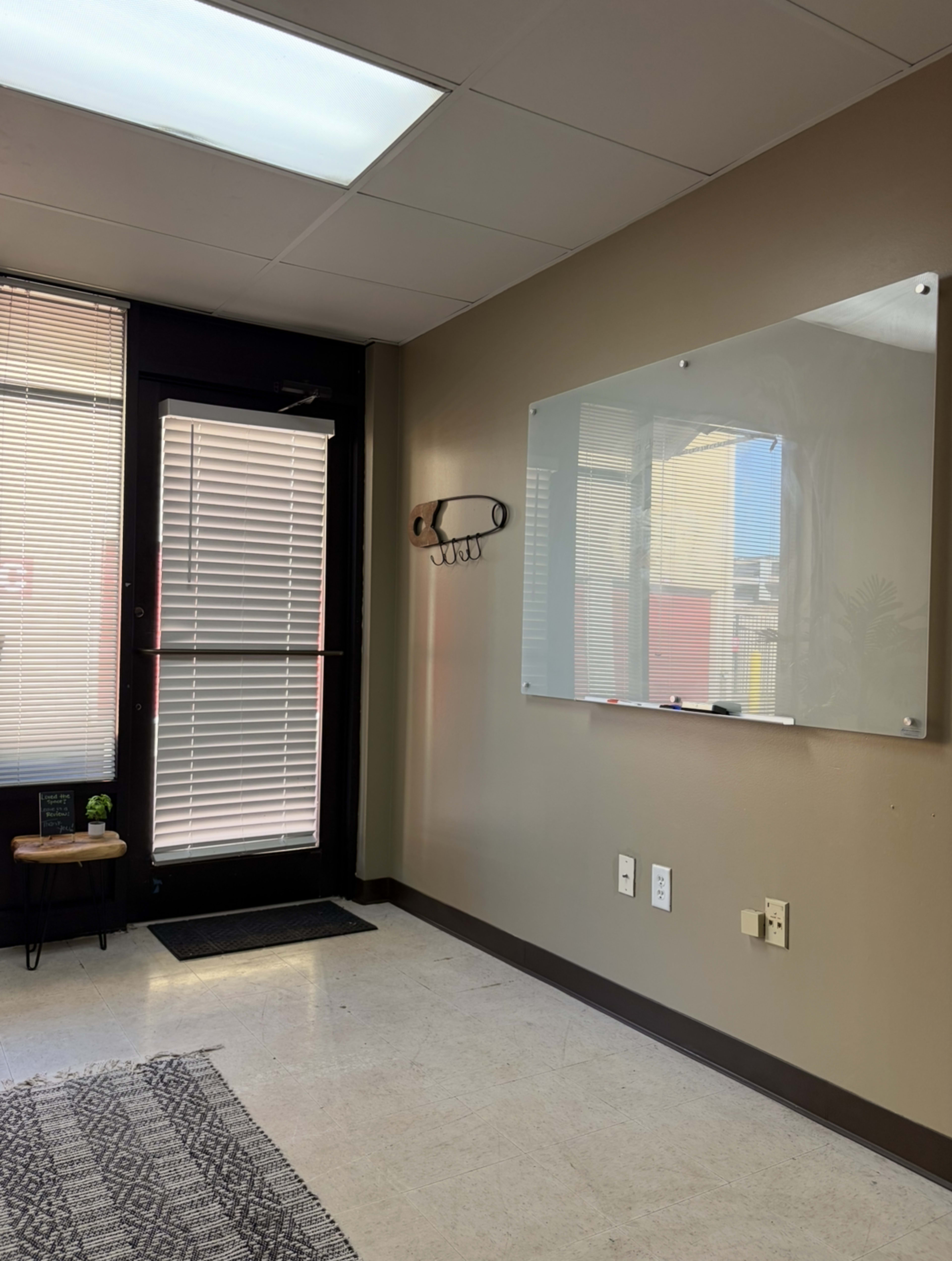 The image shows an office space with a whiteboard mounted on a beige wall, a door with blinds, and a small seating area with a rug.