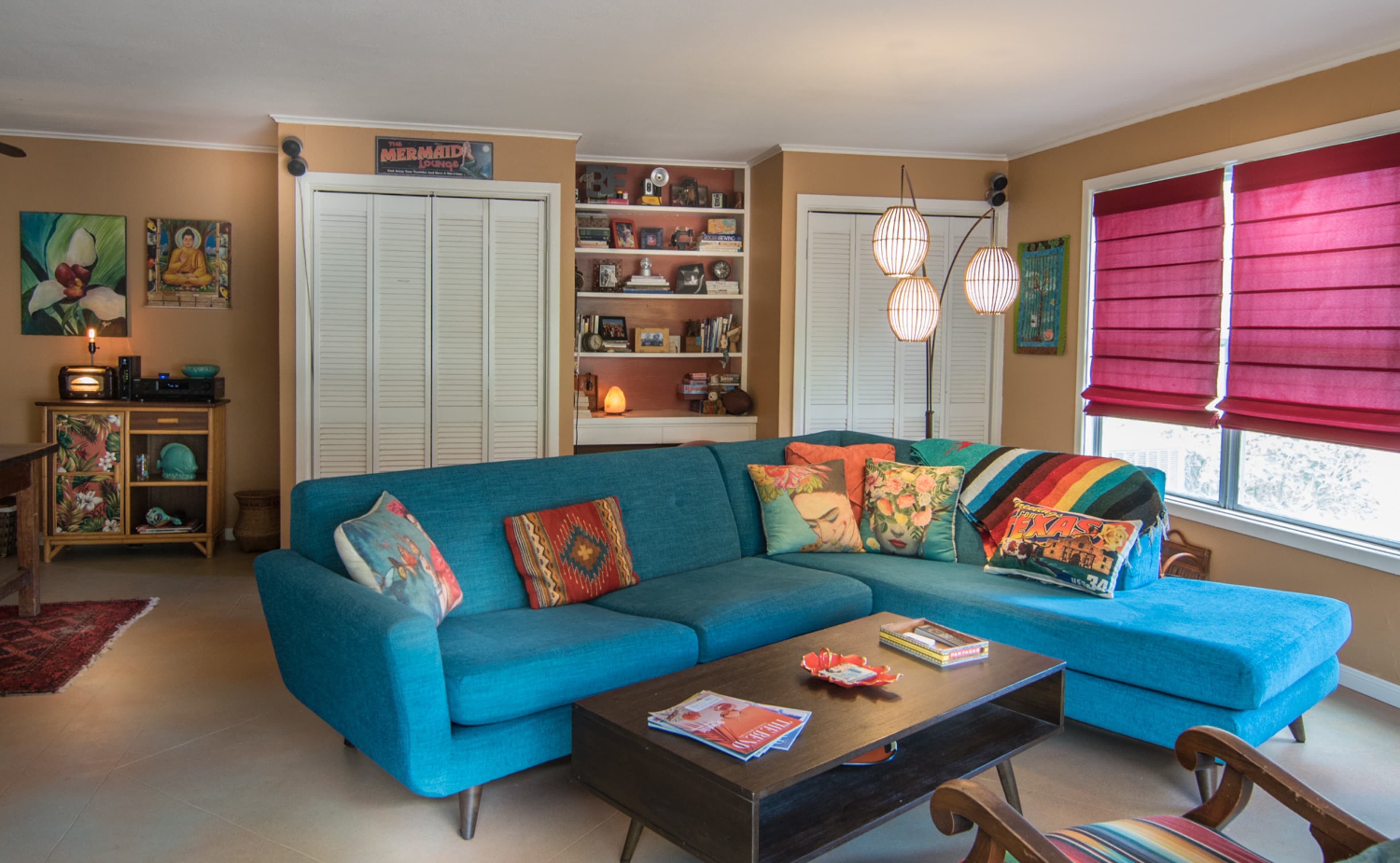 The living room features a turquoise sectional sofa adorned with colorful pillows, a dark coffee table, and large windows with red shades.