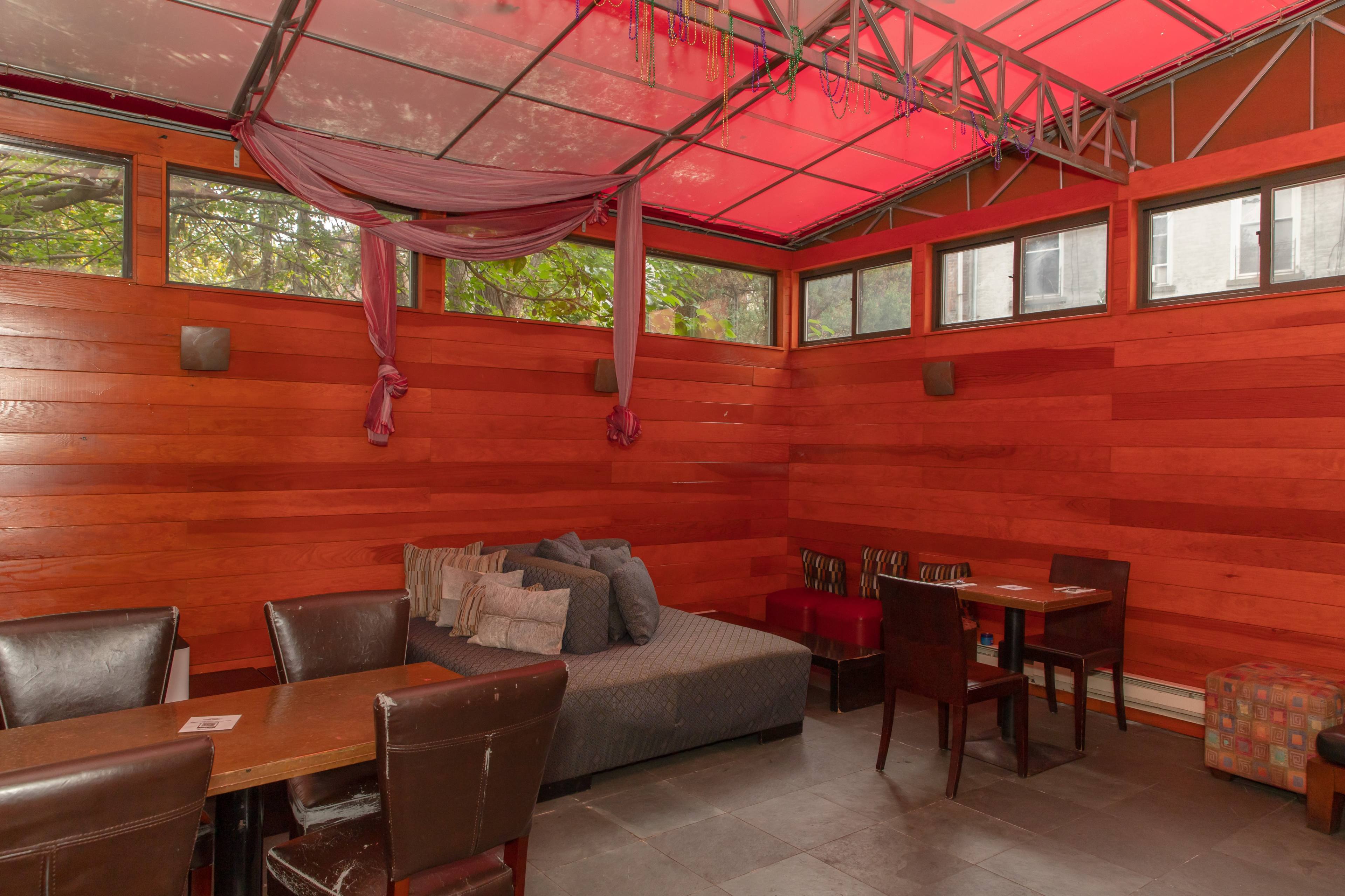 A cozy seating area with wooden walls, a large upholstered bench with pillows, and several tables and chairs under a partially transparent roof.