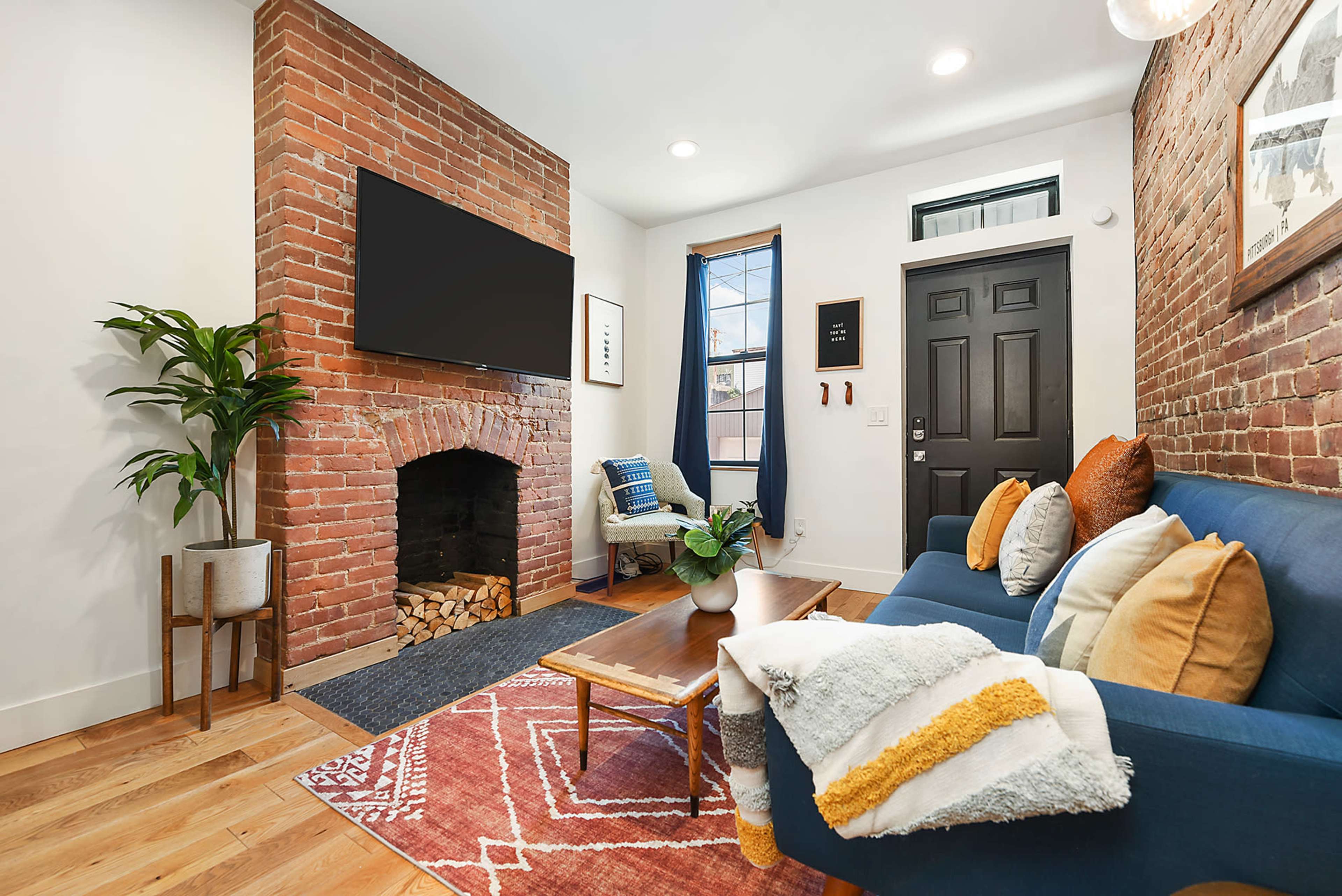 The living room features a brick accent wall, a large flat-screen TV mounted above a fireplace, and a blue sofa adorned with colorful cushions.