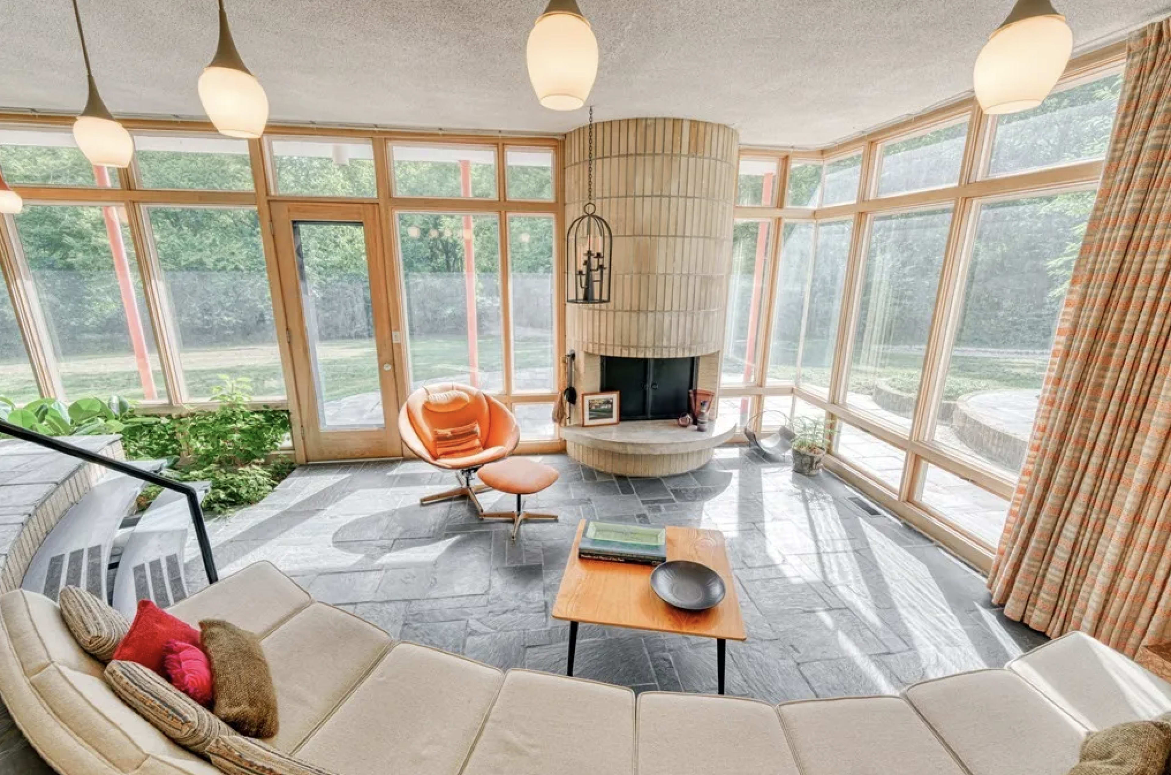 The scene features a modern living room with large windows, a circular stone fireplace at the center, a curved sofa, an orange lounge chair, and a small wooden coffee table.