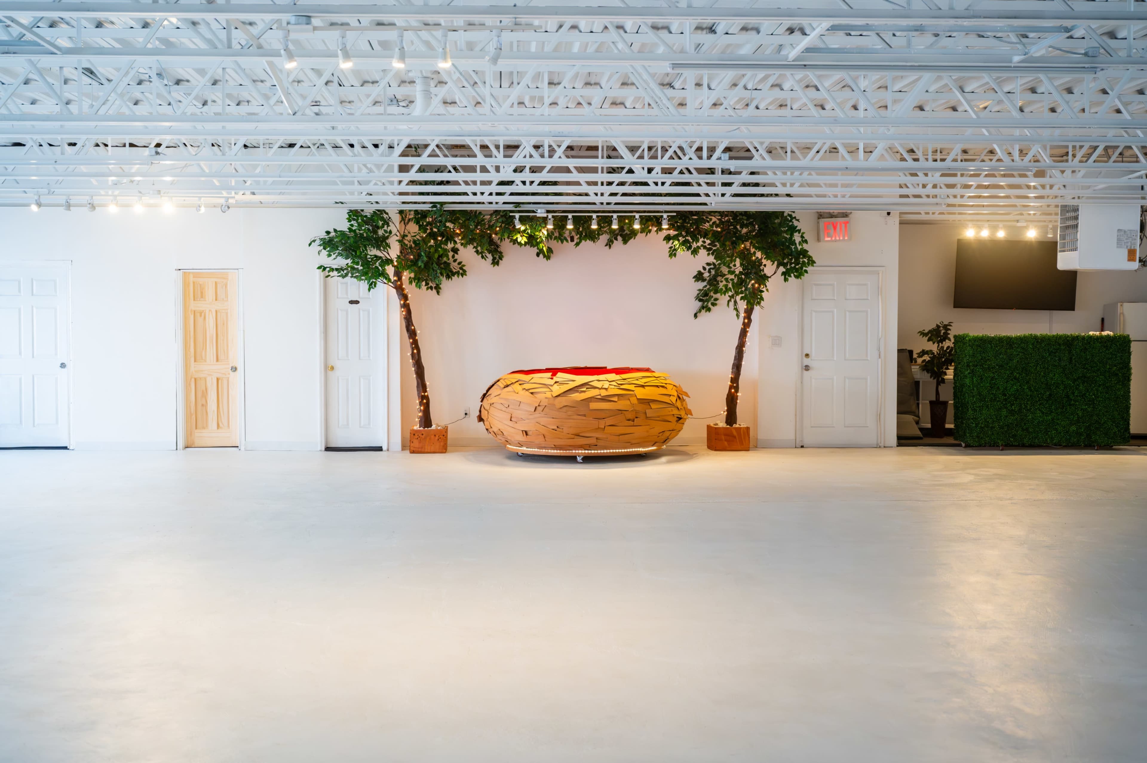 The image features a spacious, well-lit room with a large, stylized wooden donut sculpture surrounded by two trees and white walls.