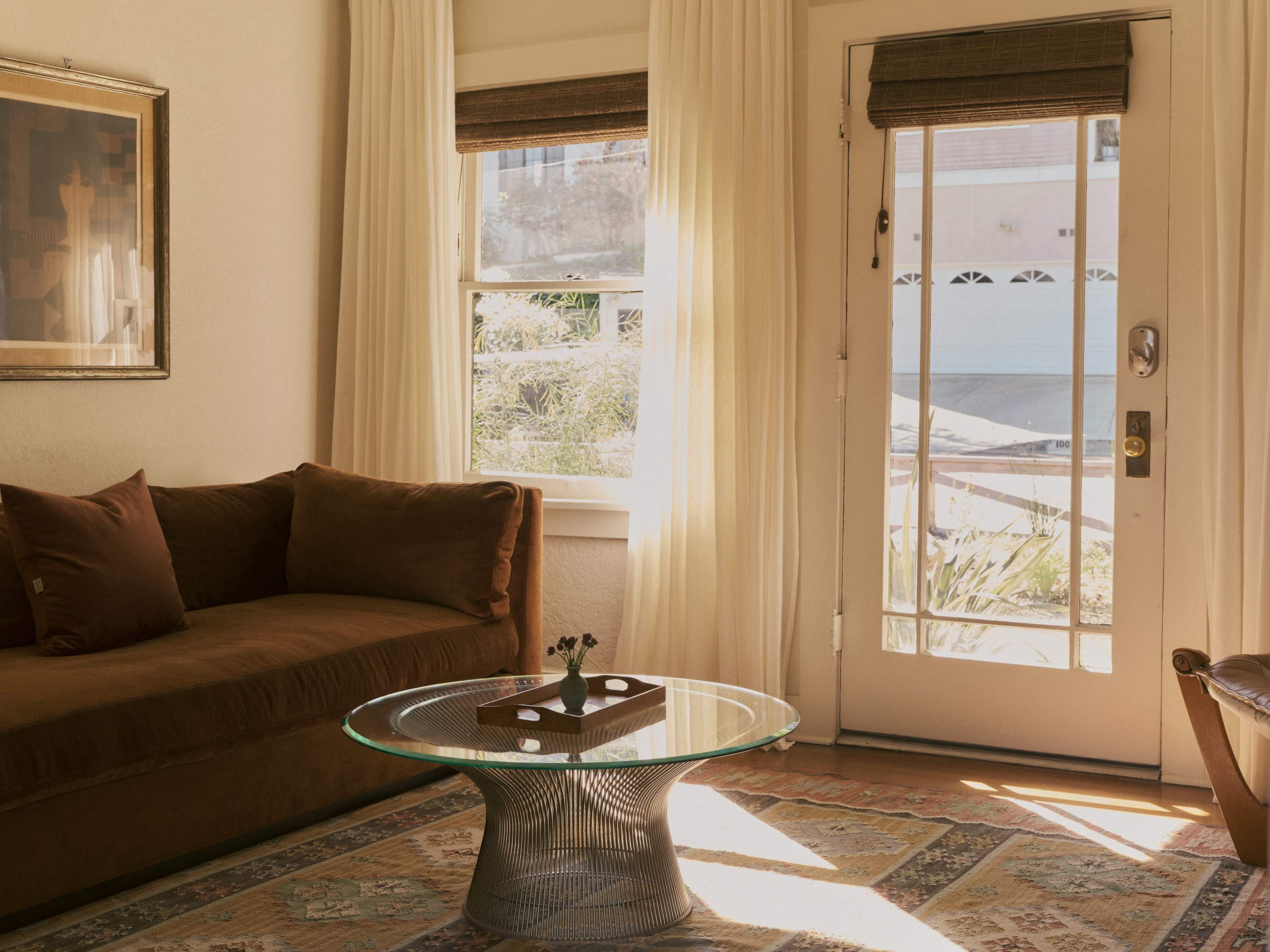 The image shows a cozy living room with a brown sofa, a glass coffee table, and sunlight streaming through a large window beside a door leading outside.