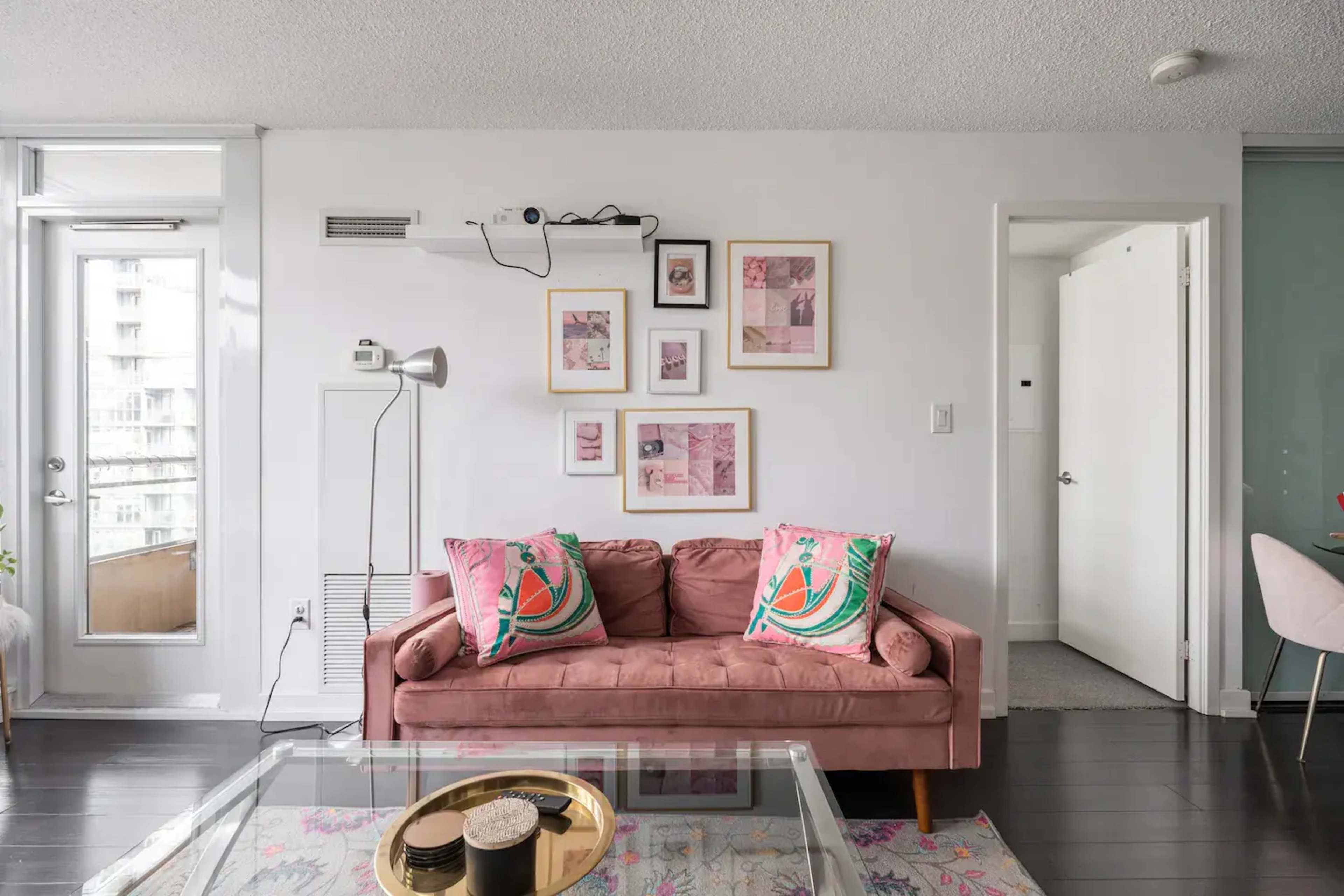 A living room features a pink sofa adorned with patterned cushions, framed artwork on the wall, and a glass coffee table.
