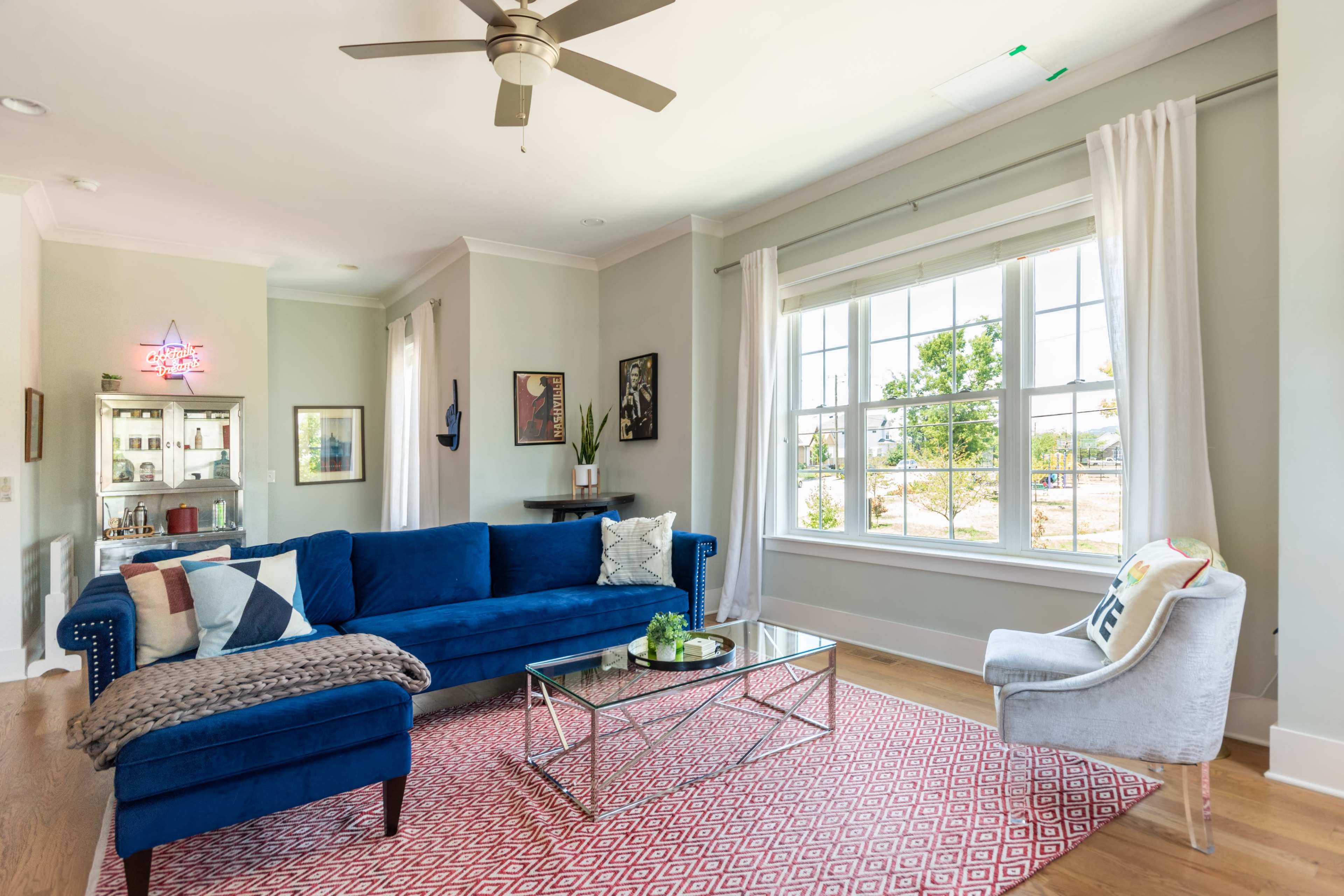 A bright living room features a blue sectional sofa, a glass coffee table, and large windows letting in natural light, with decorative artwork on the walls.