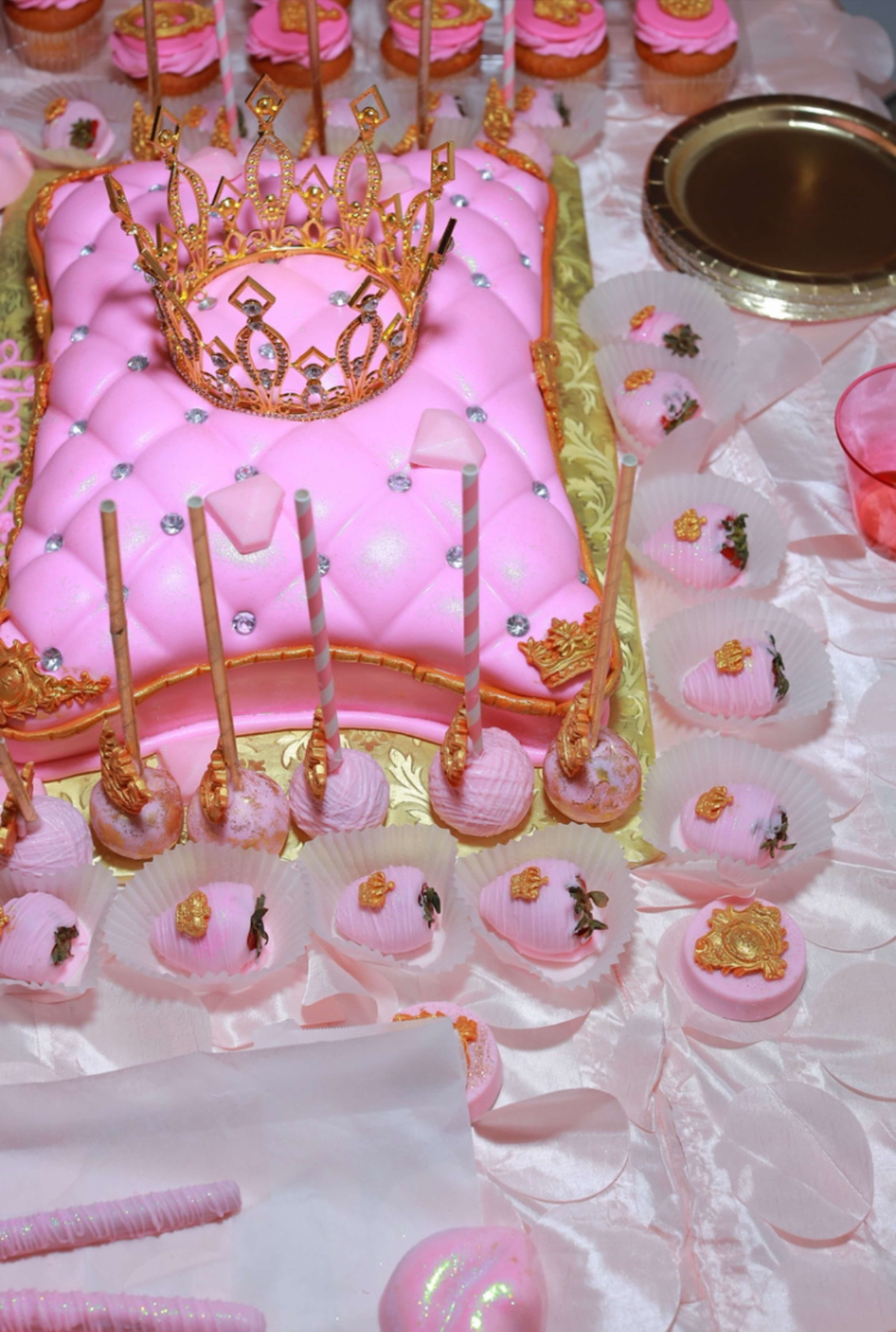 The image features a pink-themed dessert table, showcasing a large pink cake adorned with a crown and various pink sweets arranged on a decorative surface.