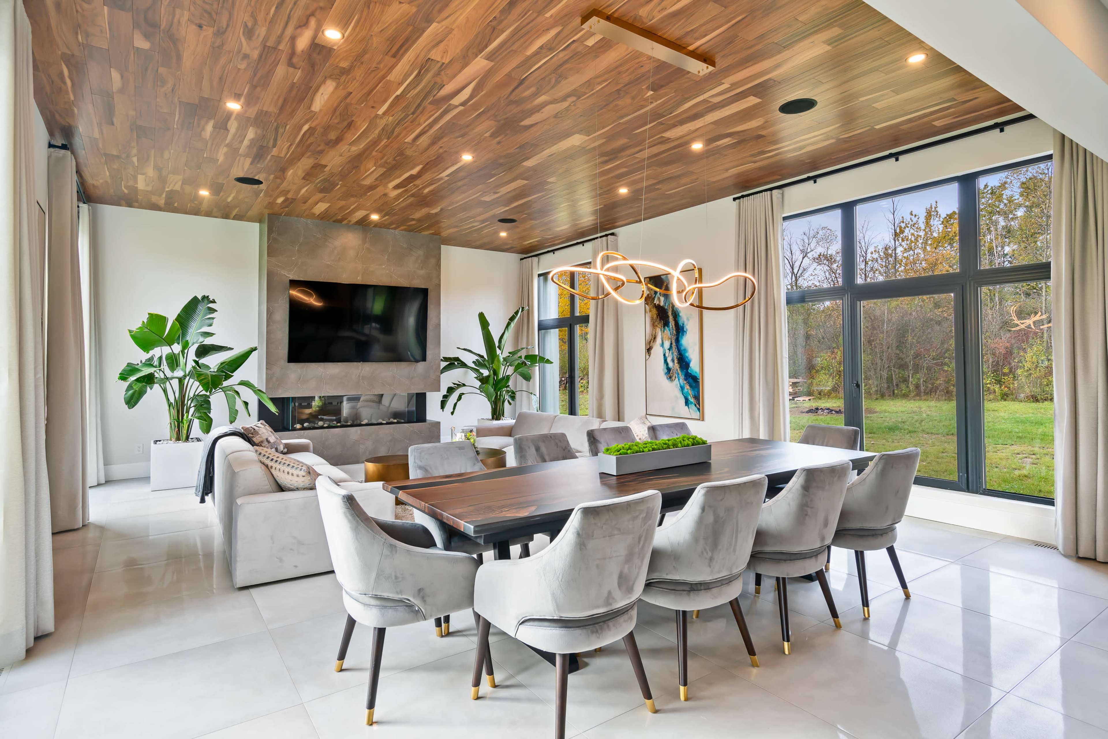 A modern dining area features a large wooden table surrounded by upholstered chairs, with a living space, fireplace, and large windows overlooking a garden.