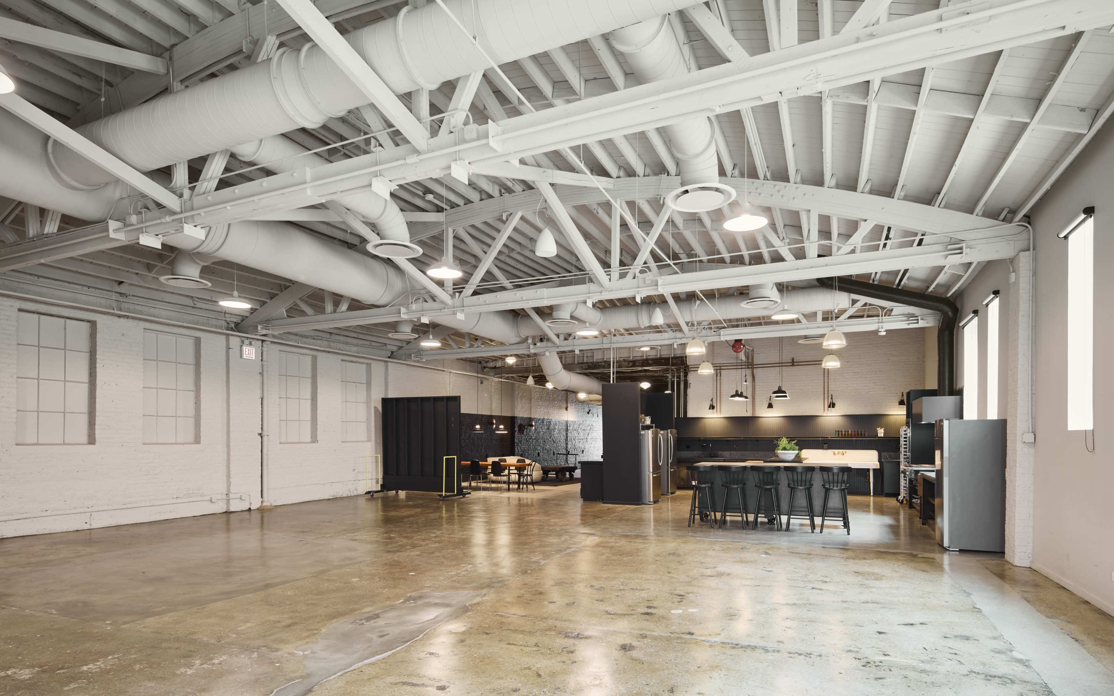 The image shows a spacious, industrial-style interior with high ceilings, exposed ductwork, and an open kitchen area equipped with modern appliances.