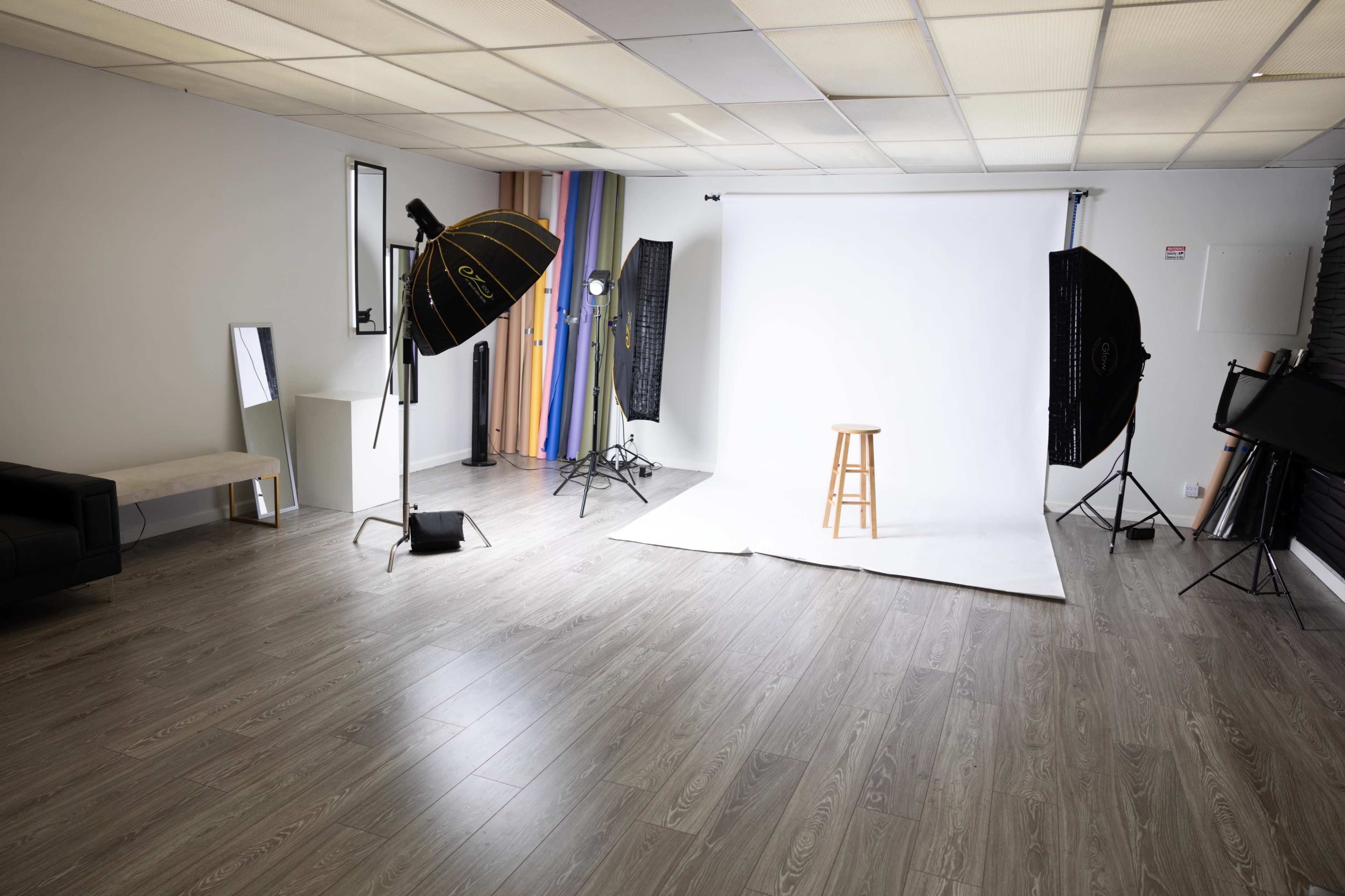 A photography studio with a white backdrop, a wooden stool, and various lighting equipment arranged around the space.